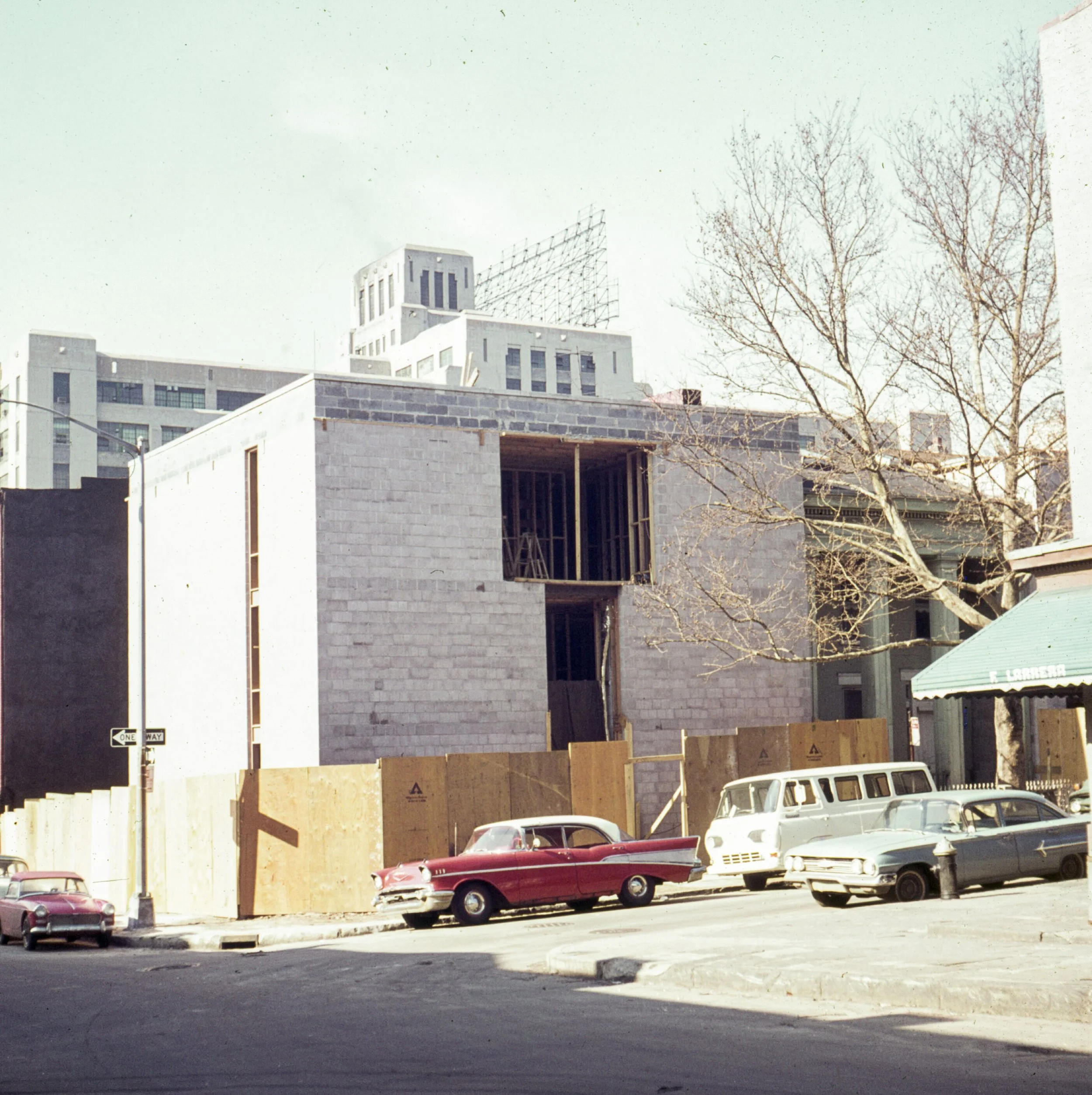 Copious cars parked along the street, with a construction site in the background featuring an unfinished building under construction, surrounded by wooden fences.