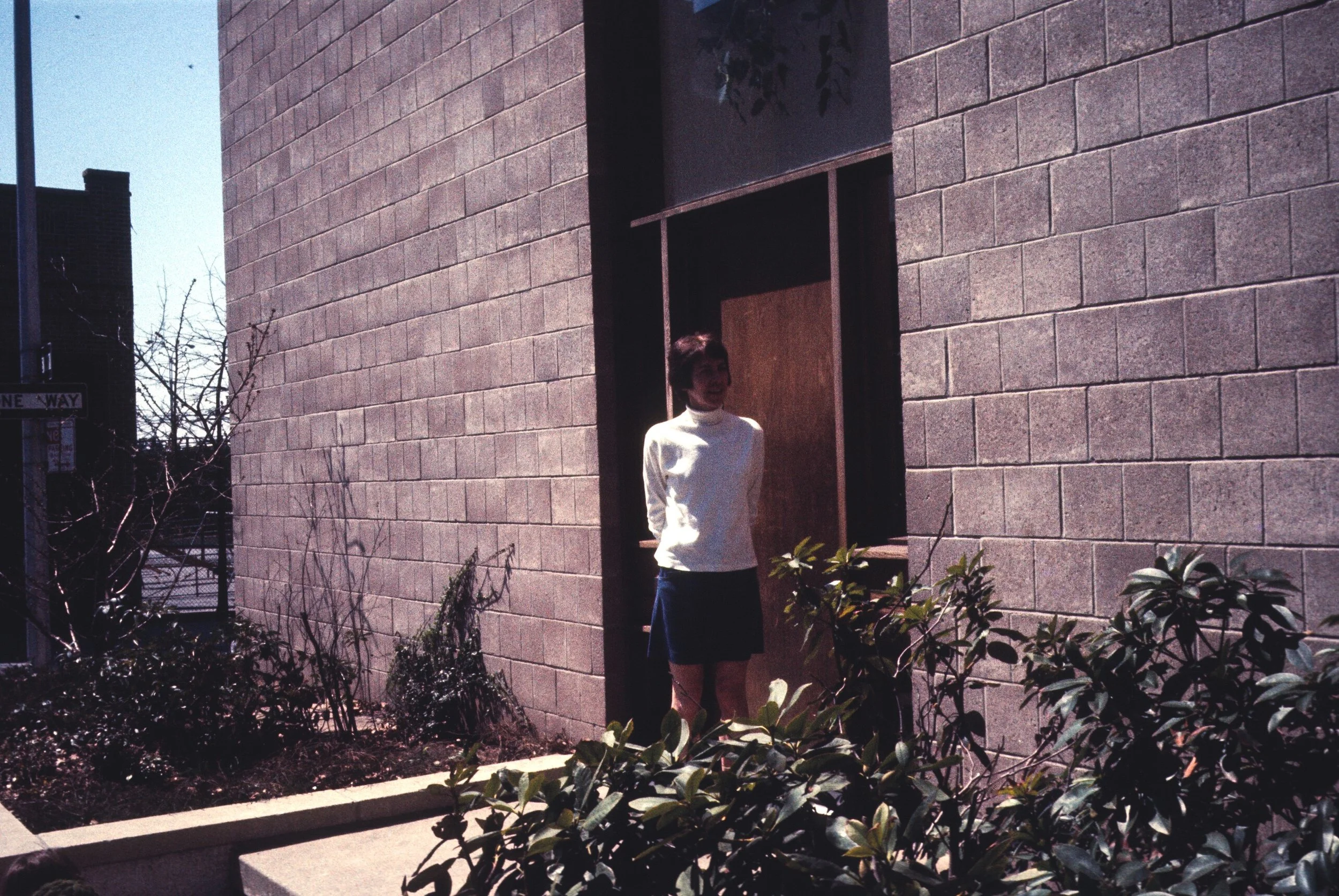 A woman with short dark hair wearing a white long-sleeve top and a dark skirt, standing outdoors next to a large brick wall with a window opening. There are plants and bushes in the foreground and a street sign in the background.