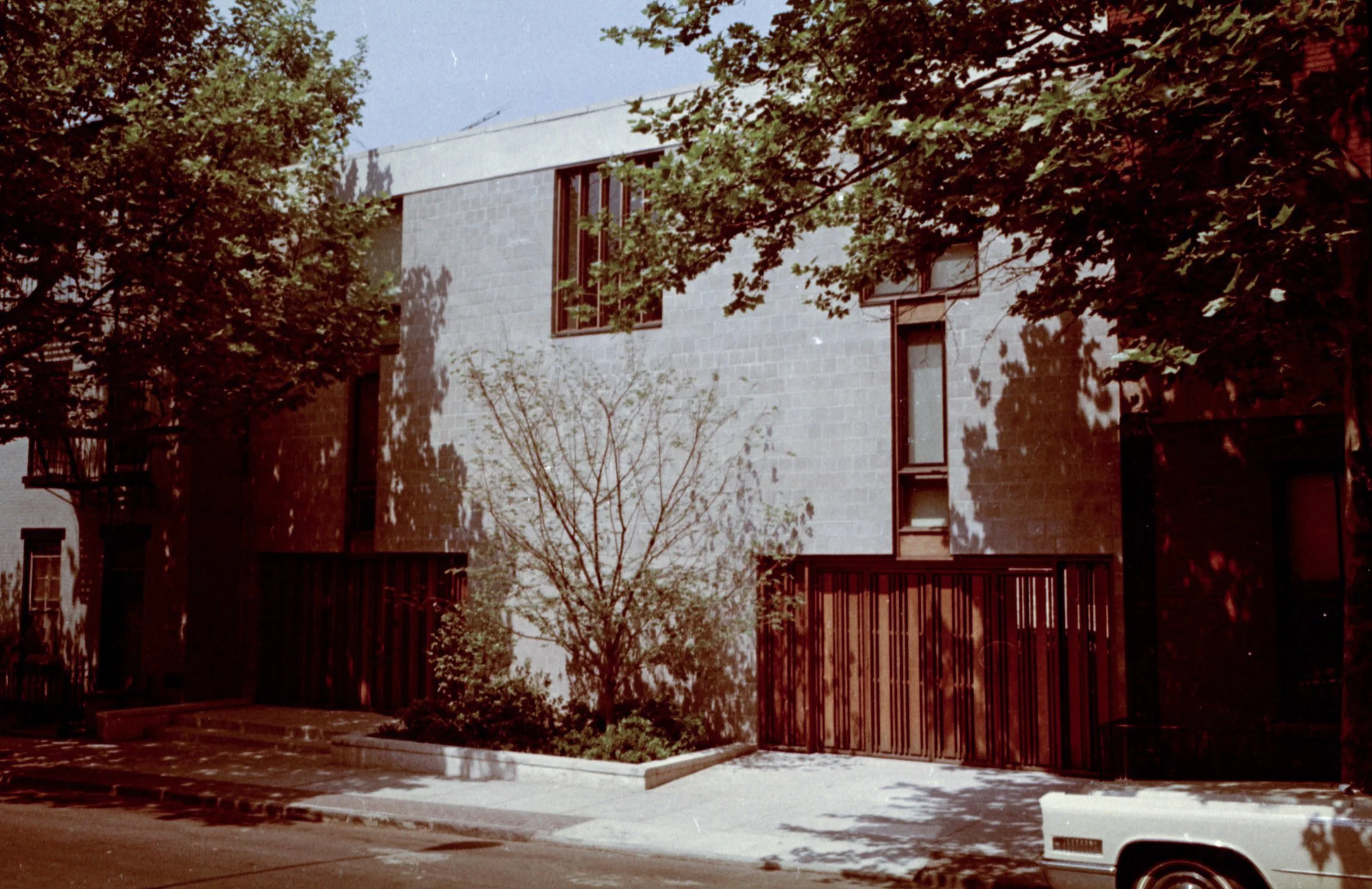 A modern residential building with large windows, surrounded by trees and a sidewalk, in a city neighborhood.