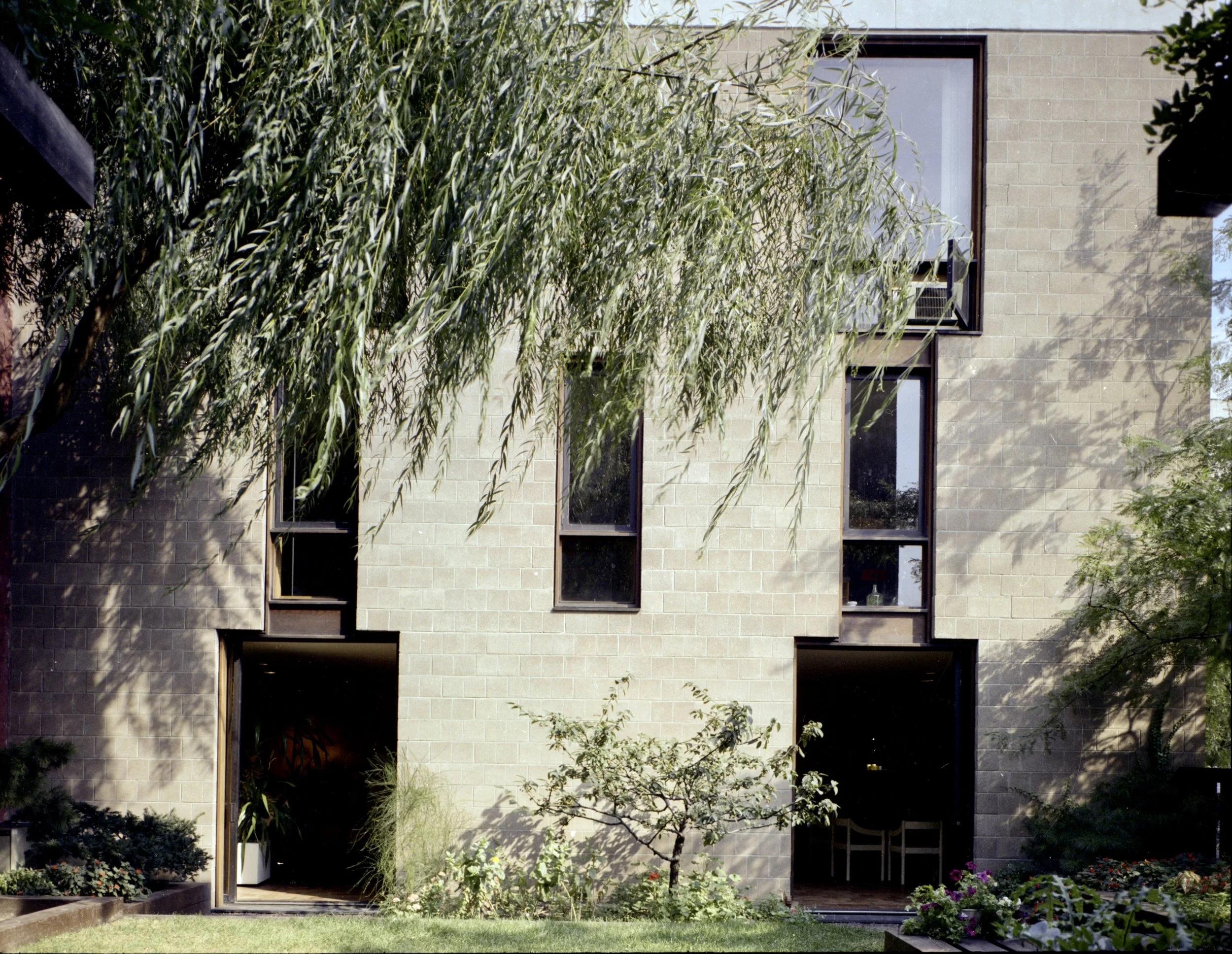 A modern building with beige brick exterior, tall windows, surrounded by trees and greenery, with a garden in the foreground.