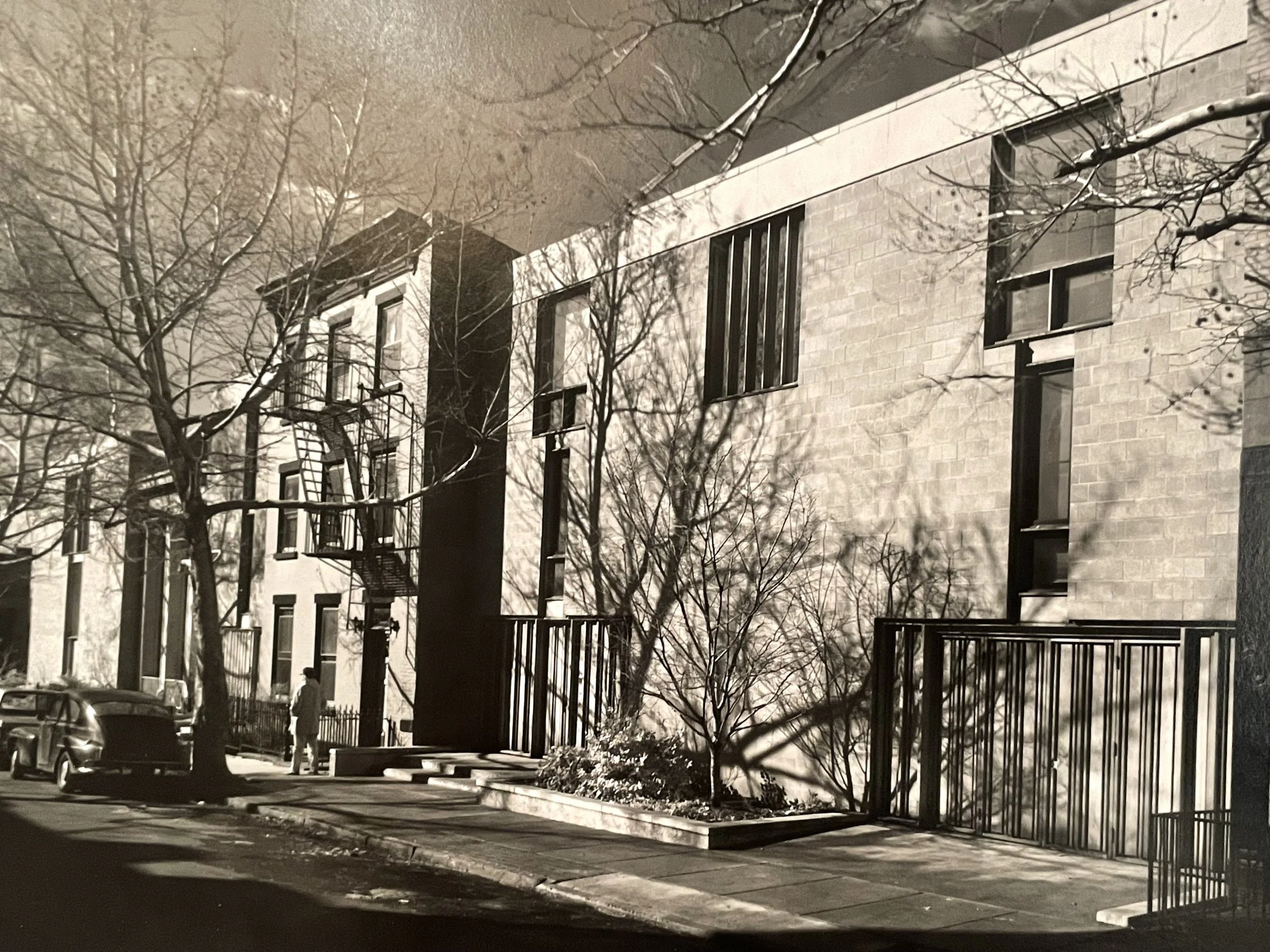 Black and white photo of a city street with a modern brick building, leafless trees, parked cars, and a person walking on the sidewalk.