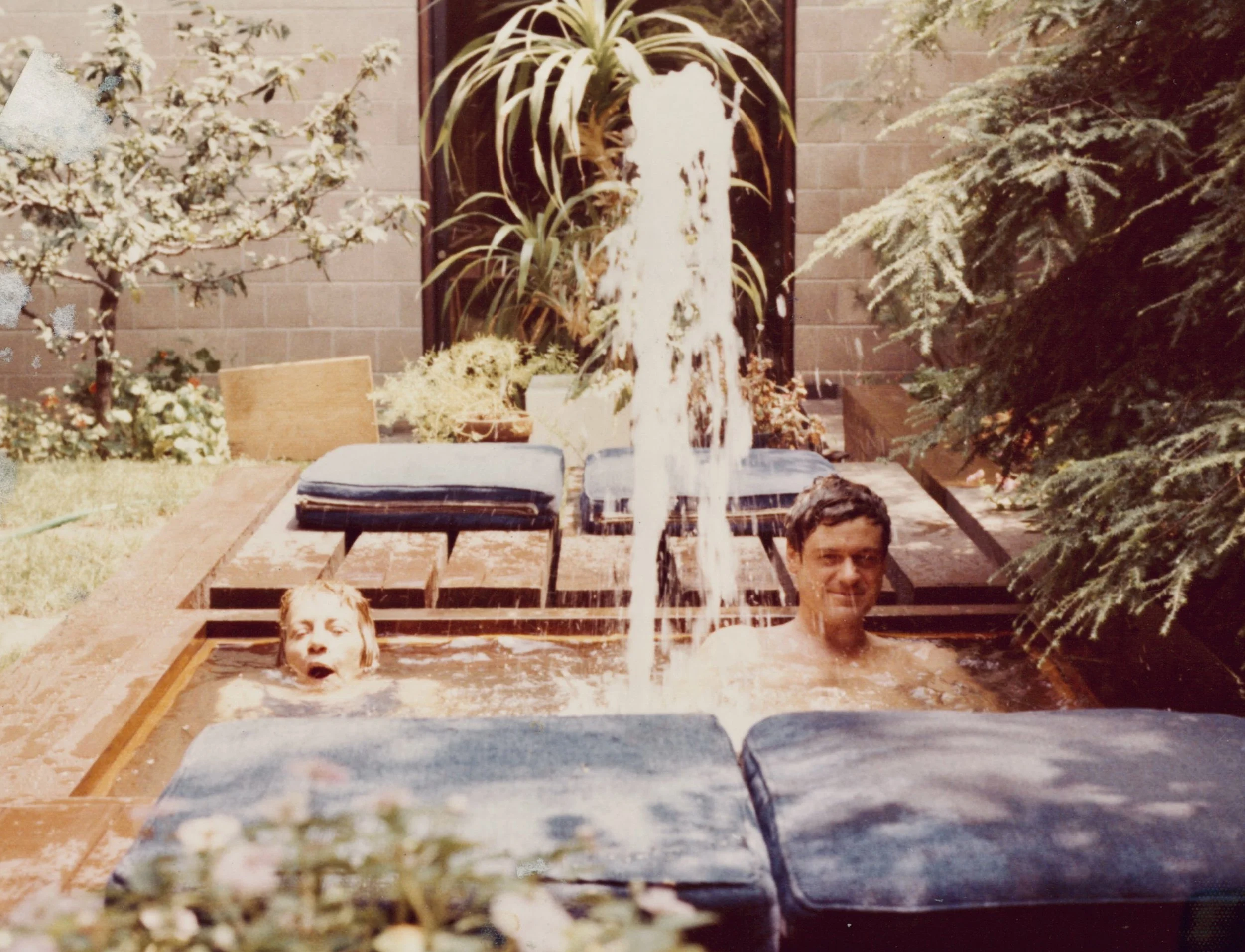 A man and a young girl enjoying time in a backyard hot tub with a cascading fountain, surrounded by plants and outdoor furniture.