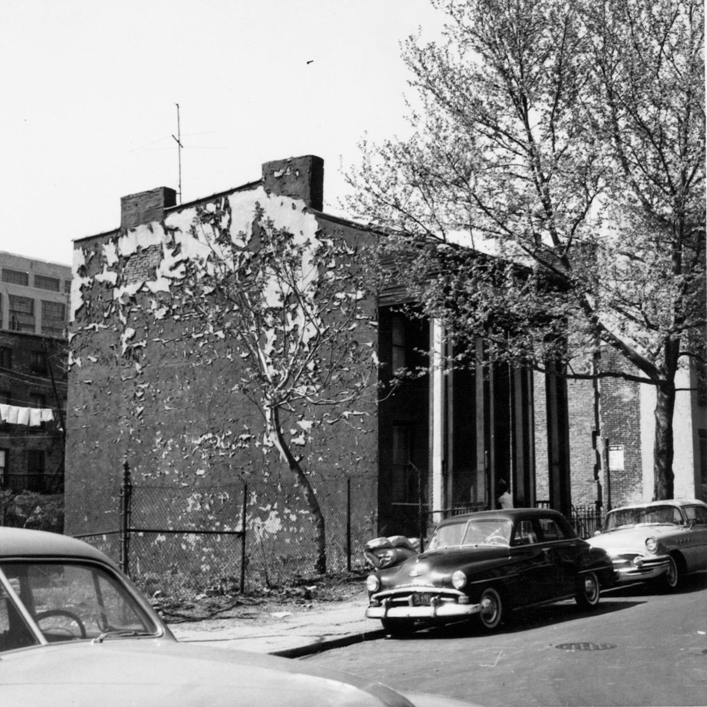 Black and white photo of an old building with peeling paint, trees, parked vintage cars, and a fenced yard in an urban setting.