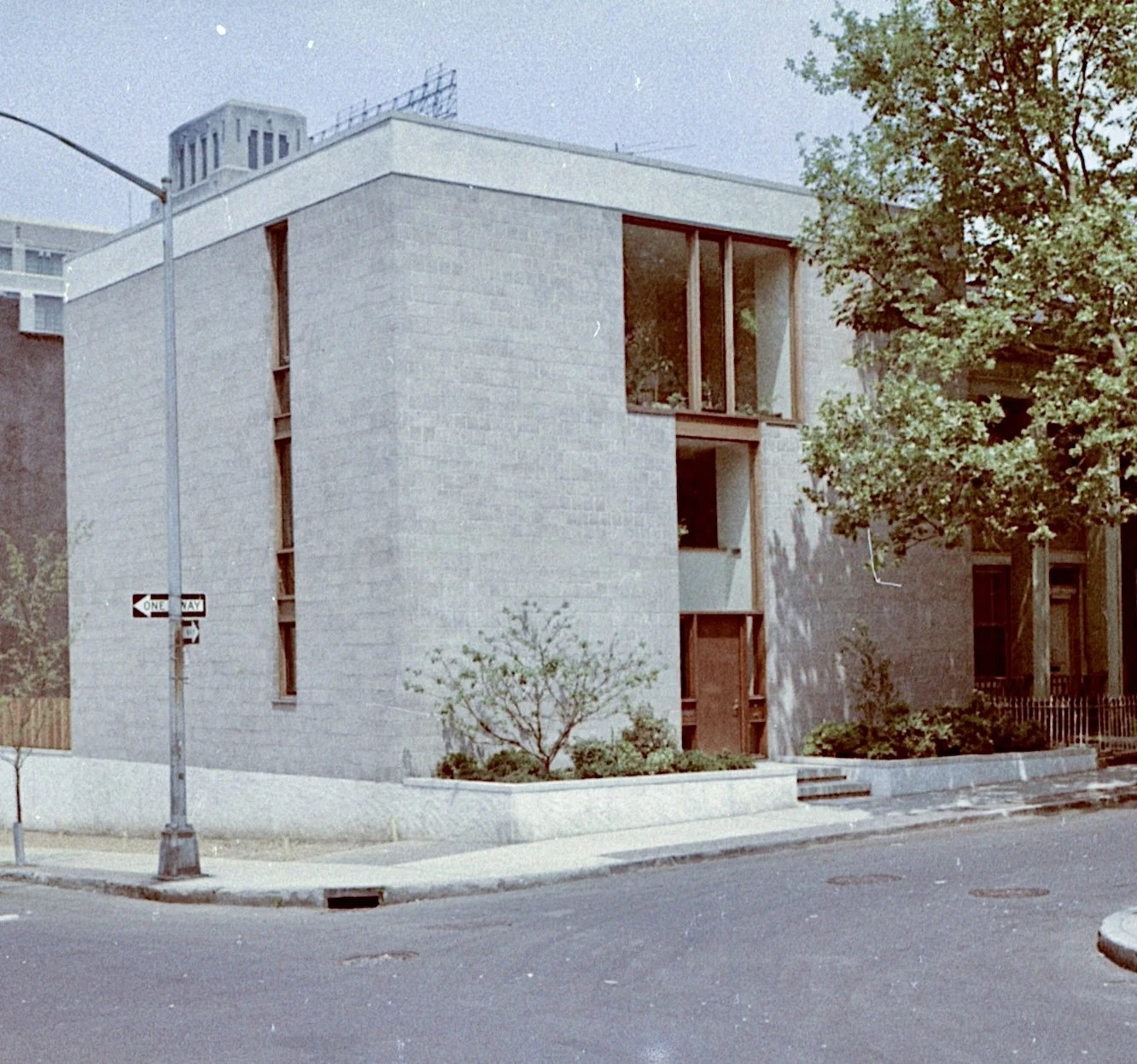 A modern residential building with light-colored brick walls, large windows, and landscaped surroundings including trees and shrubs. There is a one-way street sign in the foreground and a sidewalk, with some steps leading to the building entrance.