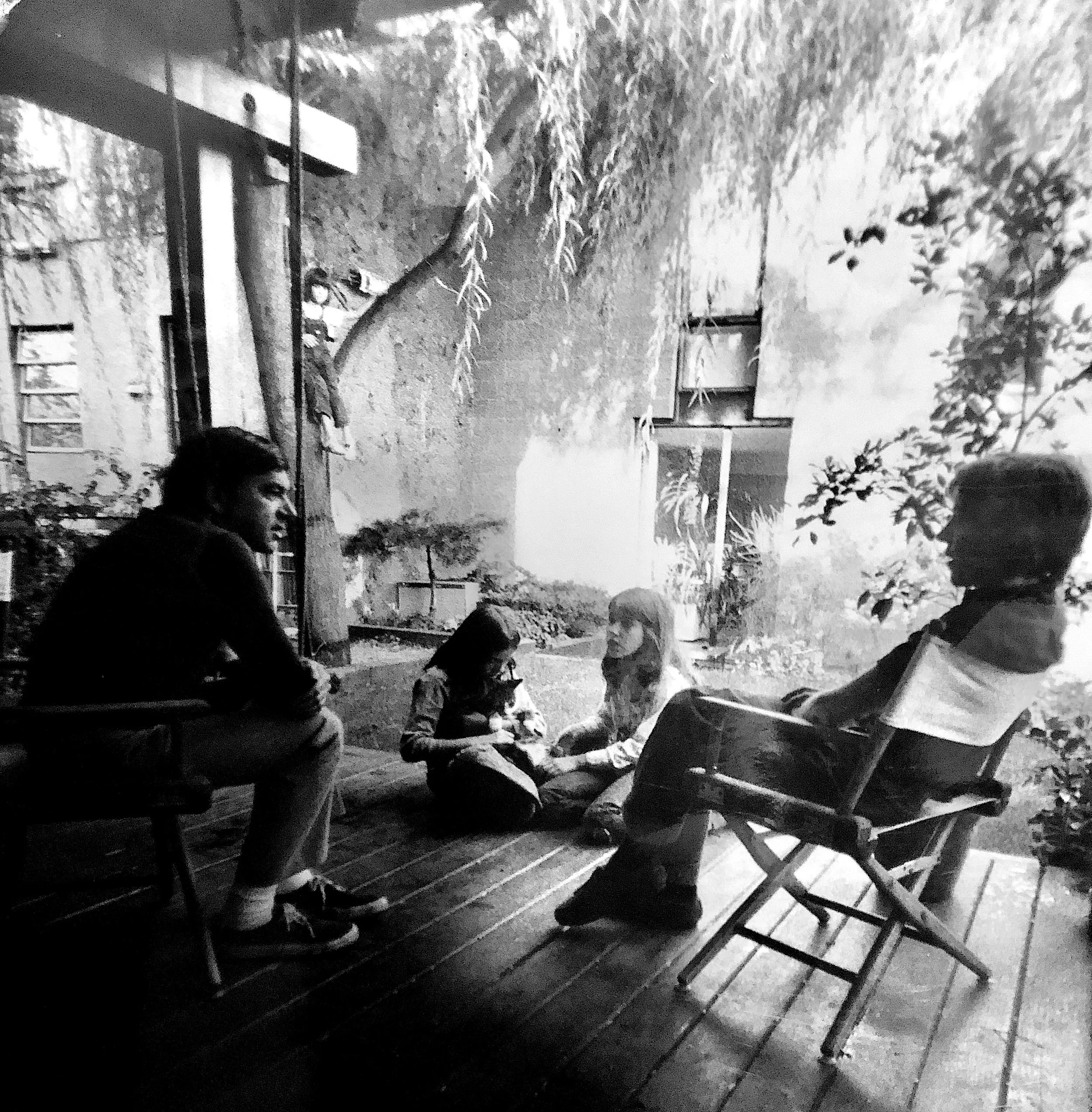 Four people sitting and engaging in conversation in a cozy outdoor setting with hanging plants and a textured wall in black and white.
