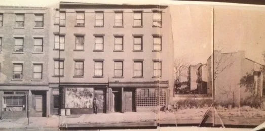 Black and white photo of a multi-story building with storefronts on the ground level, and open area with trees and a fence in the background.