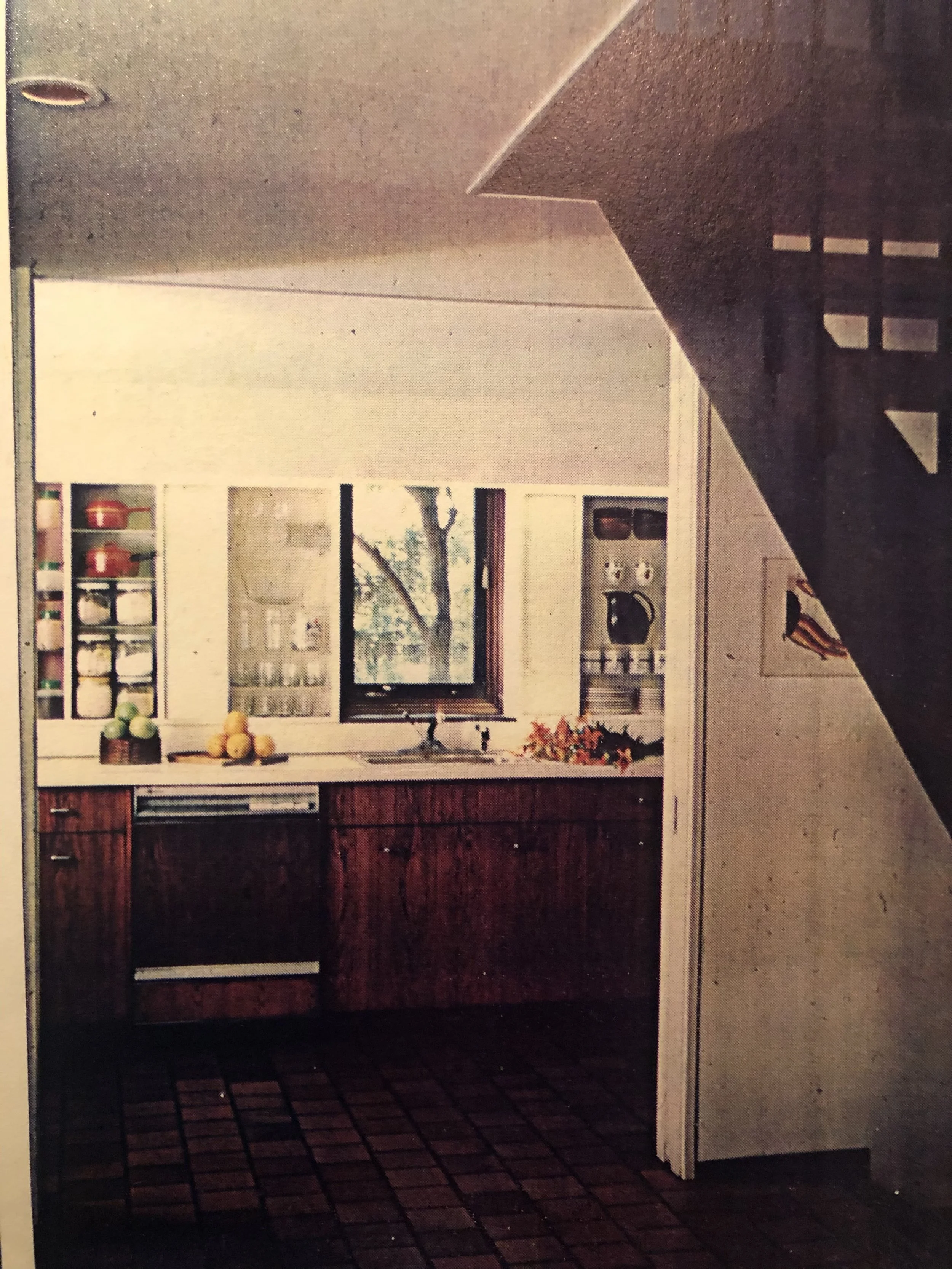 A cozy kitchen with wooden cabinets, a window overlooking trees, and a countertop with decorative items including bowls of fruit. Part of a staircase is visible on the right.