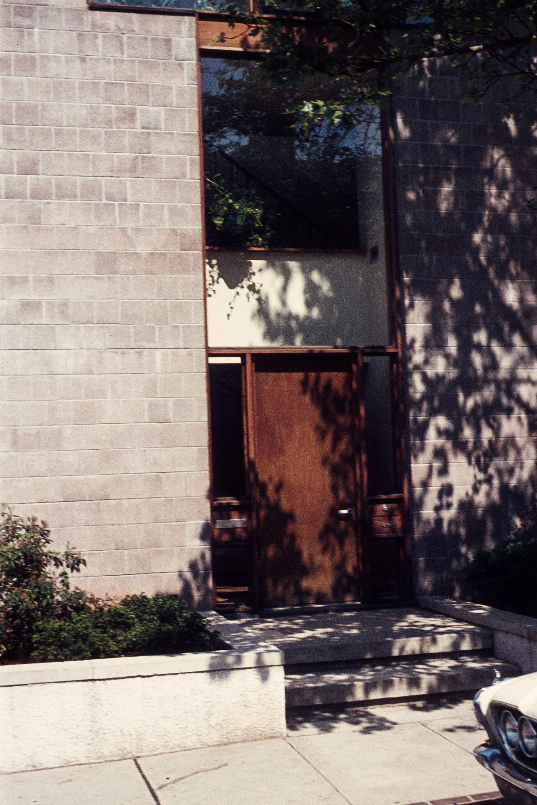 A modern building with a wooden door, concrete steps, and a window, partly shaded by tree branches, with a vintage car visible on the right side of the image.