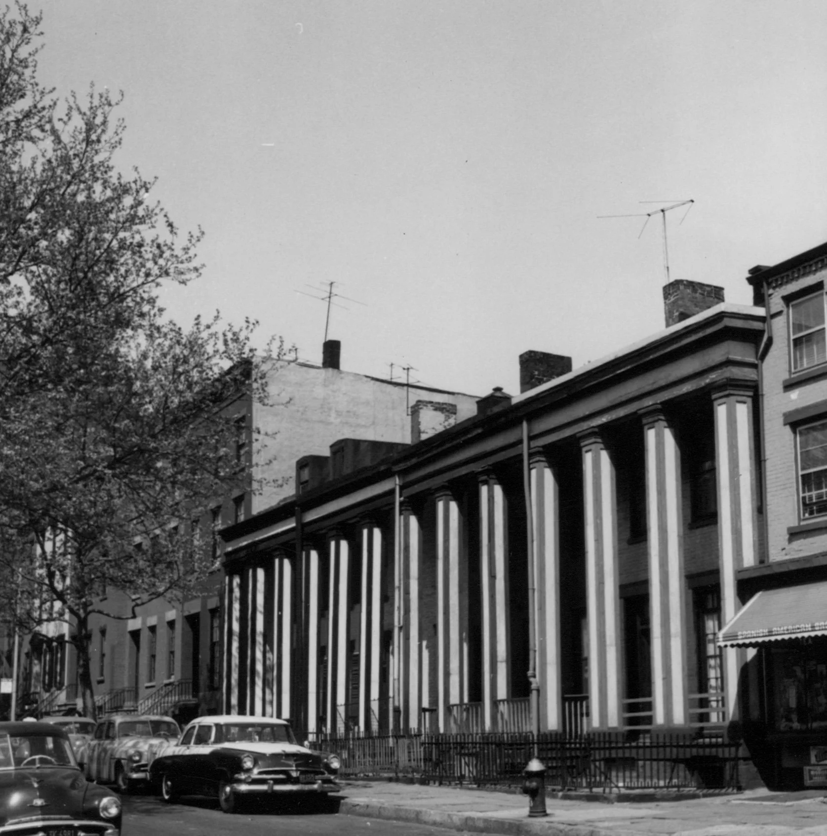 Black and white photo of a historic building with tall columns on a city street, with cars parked along the curb and trees on the sidewalk.