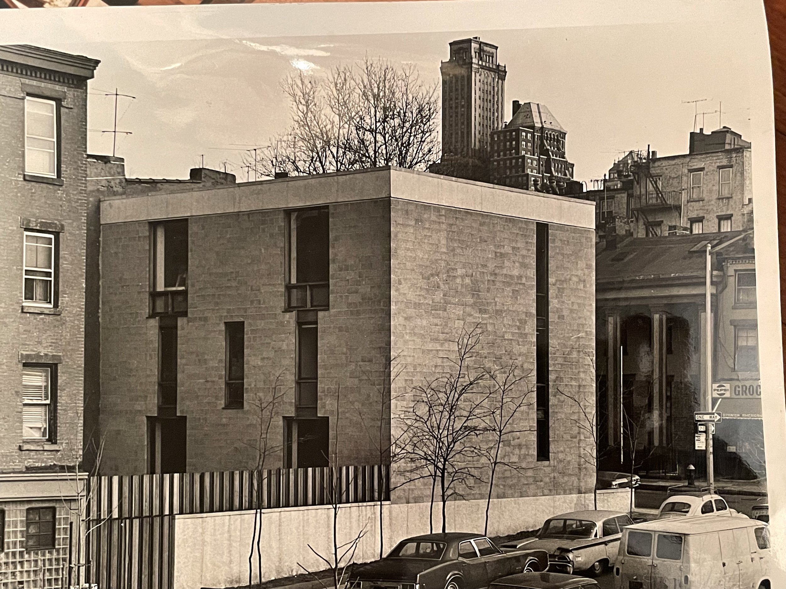 Black and white photo of a modern, rectangular building with narrow vertical windows and a flat roof, surrounded by older buildings and parked cars, with leafless trees in the foreground and a city skyline in the background.