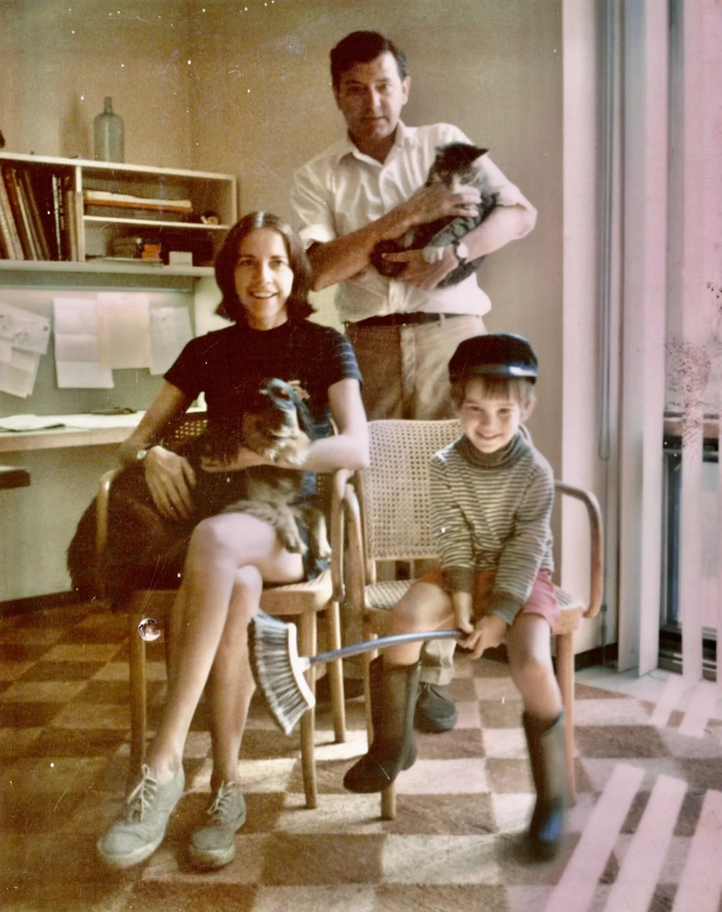 A family of four with two children, a girl and a boy, and their two dogs. The girl is sitting on a chair holding a small dog, the boy is sitting on the floor with a broom, and the dad is standing behind them holding a dog. The family is in a room wit