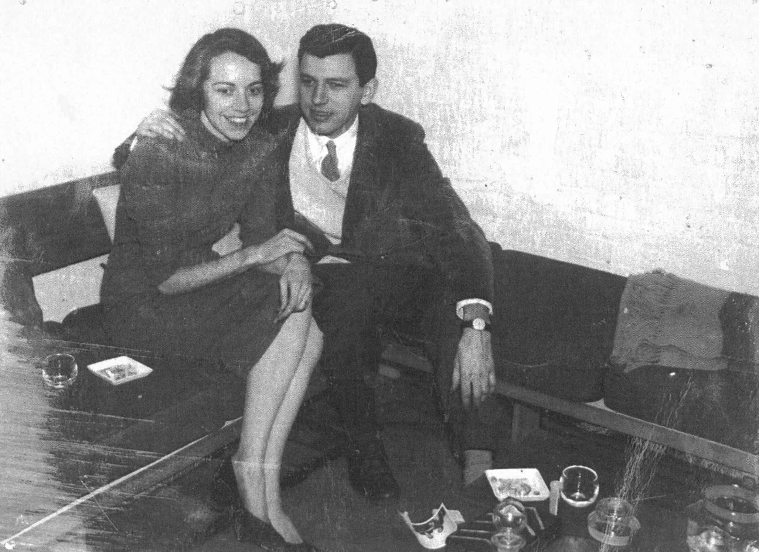 A vintage black and white photograph of a woman and a man sitting on a sofa, smiling and leaning towards each other. There are drinks, a cigarette, and small plates on the table in front of them.