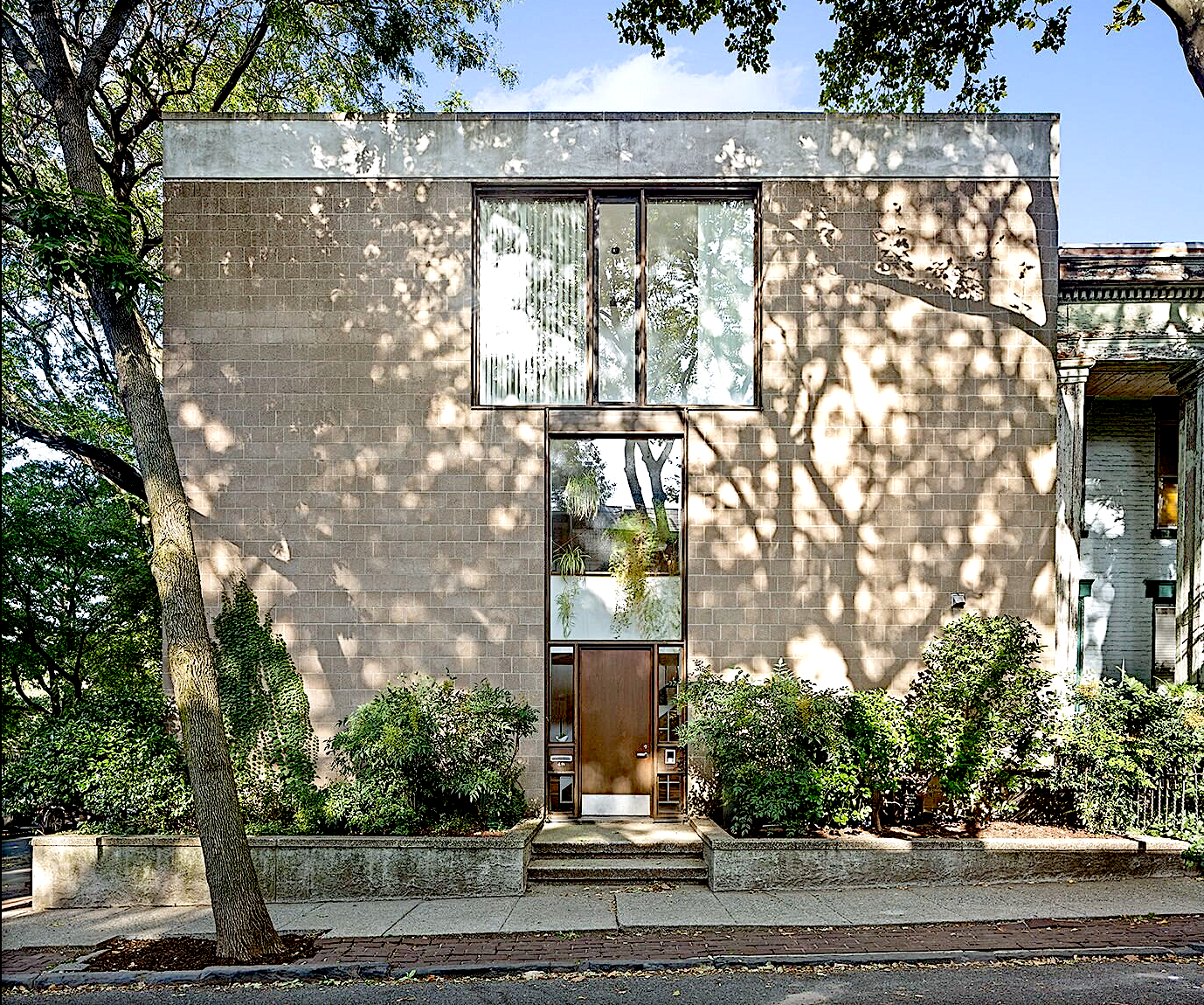 A modern, two-story residential building with a brick facade, surrounded by greenery and casting dappled shadows from trees. It has a central wooden door, large windows above and beside it, and is enclosed by a concrete planter with bushes.