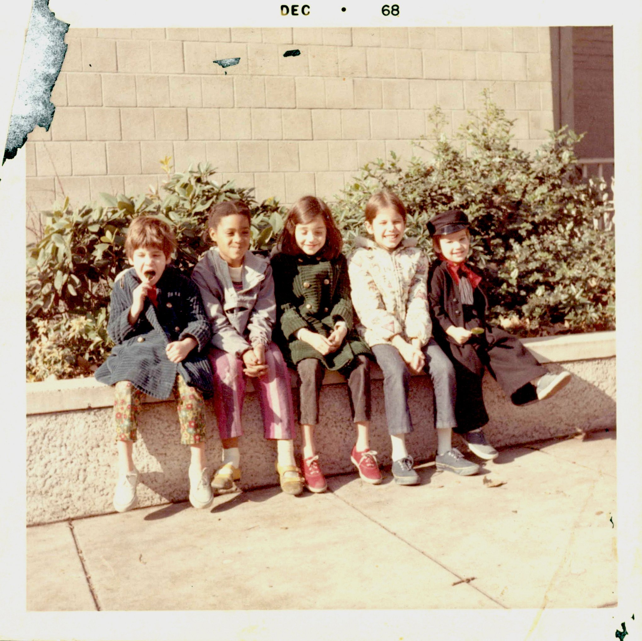 Five children sitting on a concrete ledge in front of bushes and a brick wall, smiling and posing for the camera. The children are wearing colorful jackets and shoes.