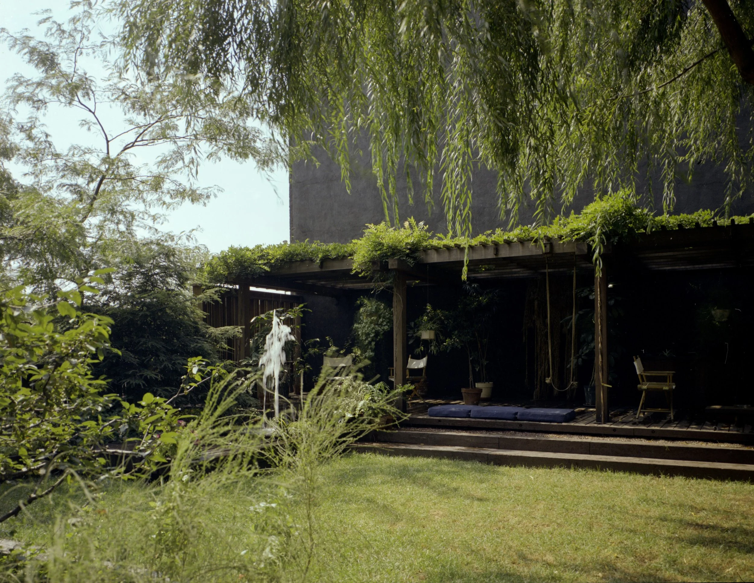 A backyard with lush green grass, a wooden deck with outdoor chairs, a blue cushion, potted plants, a water fountain, and a shaded roof with greenery hanging from trees.