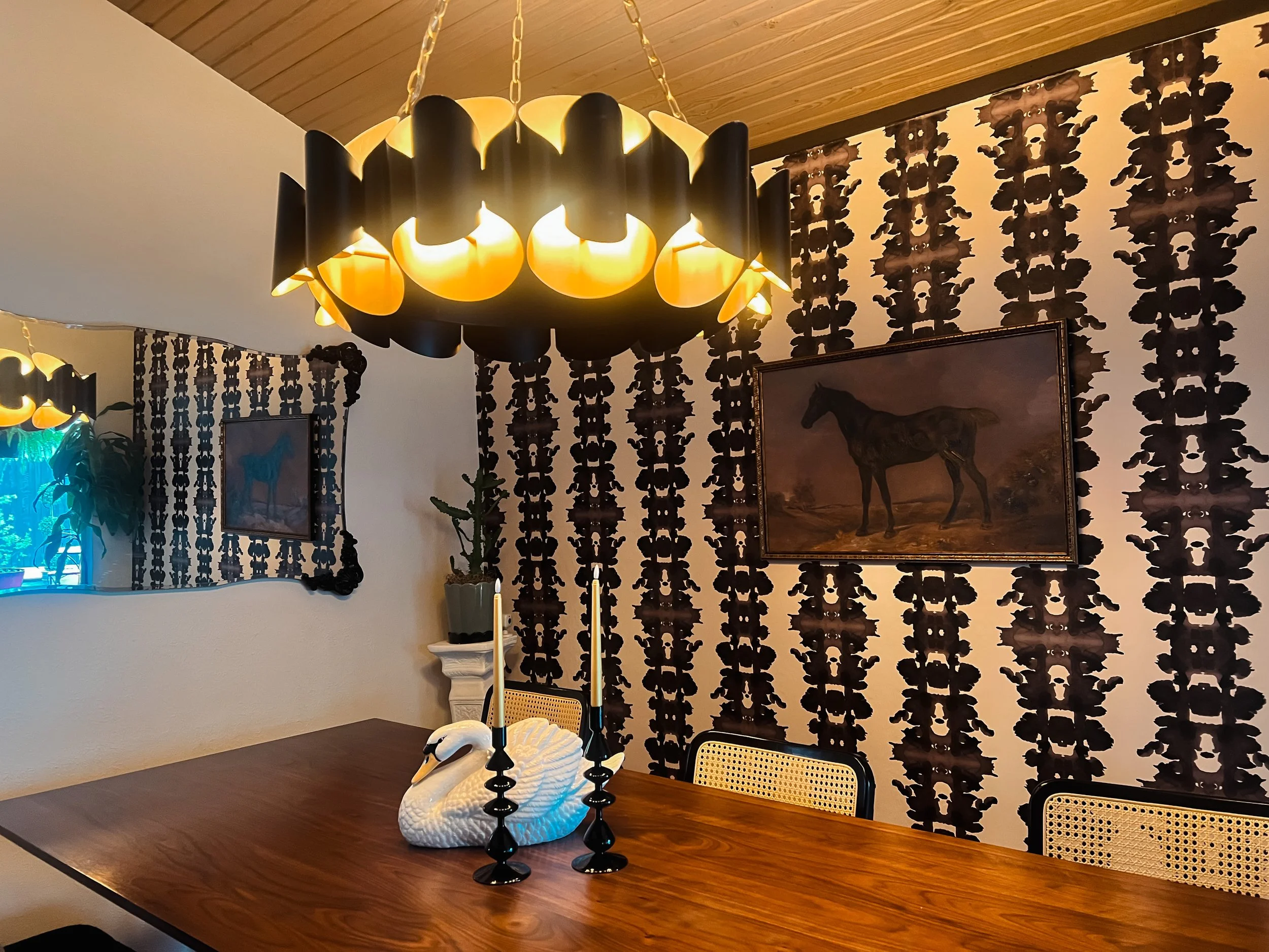 A dining room with a wooden table decorated with a white swan sculpture and black candle holders, a black and yellow chandelier, patterned wallpaper with black floral designs, a painting of a galloping horse, and a mirror with a black ornate frame.