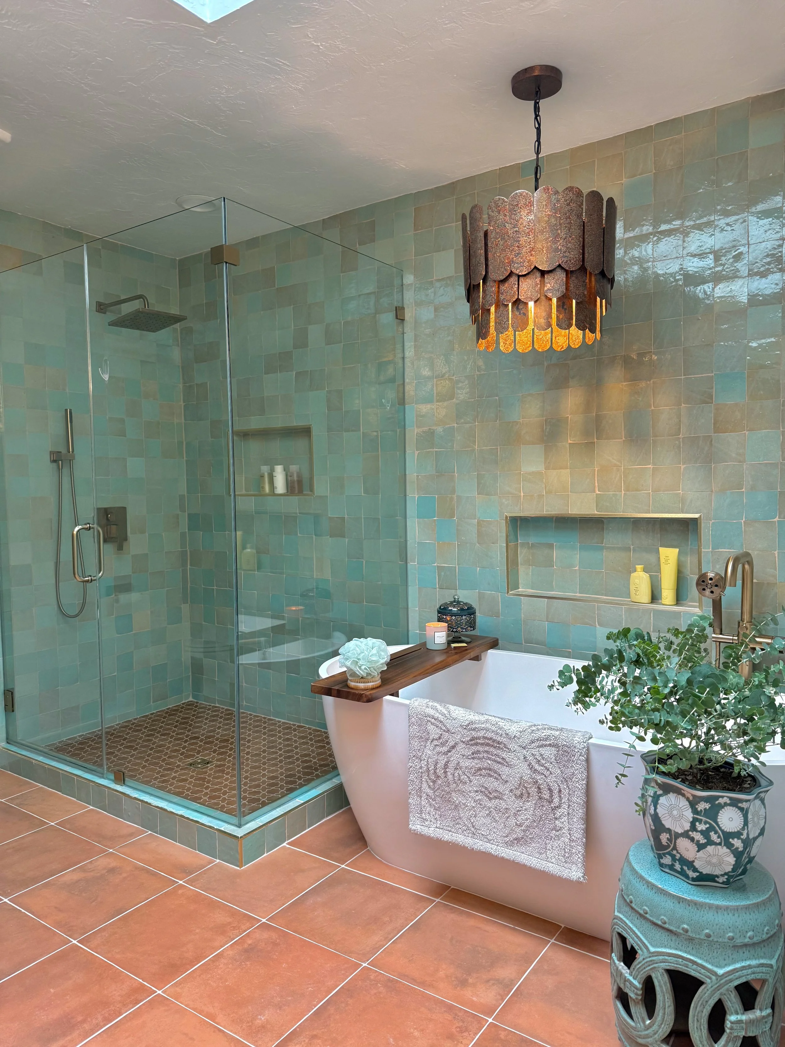 A bathroom with a glass-enclosed shower area, a bathtub with a towel hanging on the side, a side table with a potted plant, and various toiletries. The bathroom has teal and beige tiled walls and terra cotta floor tiles. A brown pendant light hangs f