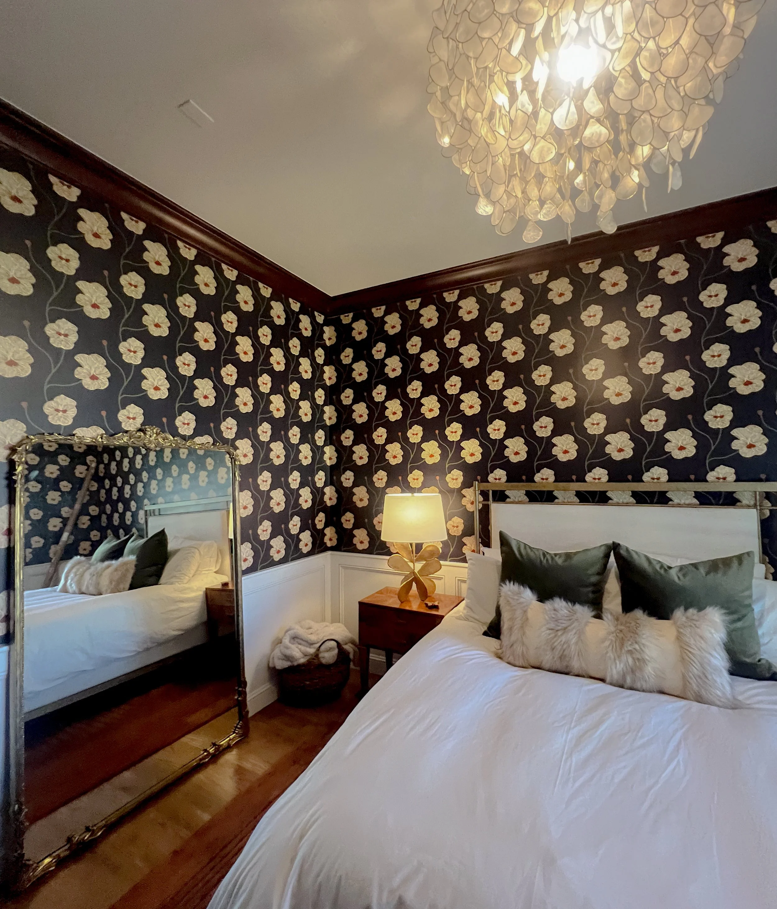 A cozy bedroom with floral wallpaper, a large bed with white bedding and decorative pillows, a wooden nightstand with a lamp, and a full-length mirror reflecting part of the bed.