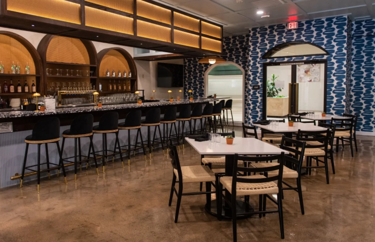 Interior of a modern restaurant or bar with a marble-topped counter, black bar stools, and small tables with black chairs. Decor includes geometric wallpaper and hanging lighting, with a doorway and plants visible.