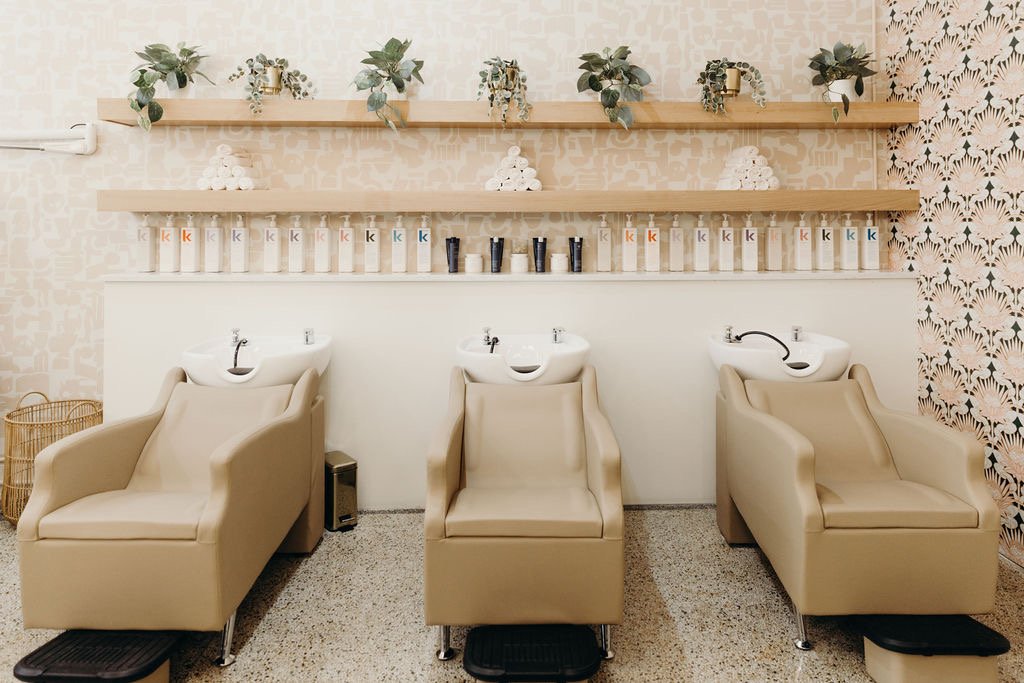 Three beige salon chairs with built-in white sinks for hair washing, set against a patterned wall. Wooden shelves above hold potted plants and hair care products, including bottles labeled with a lowercase 'k'.