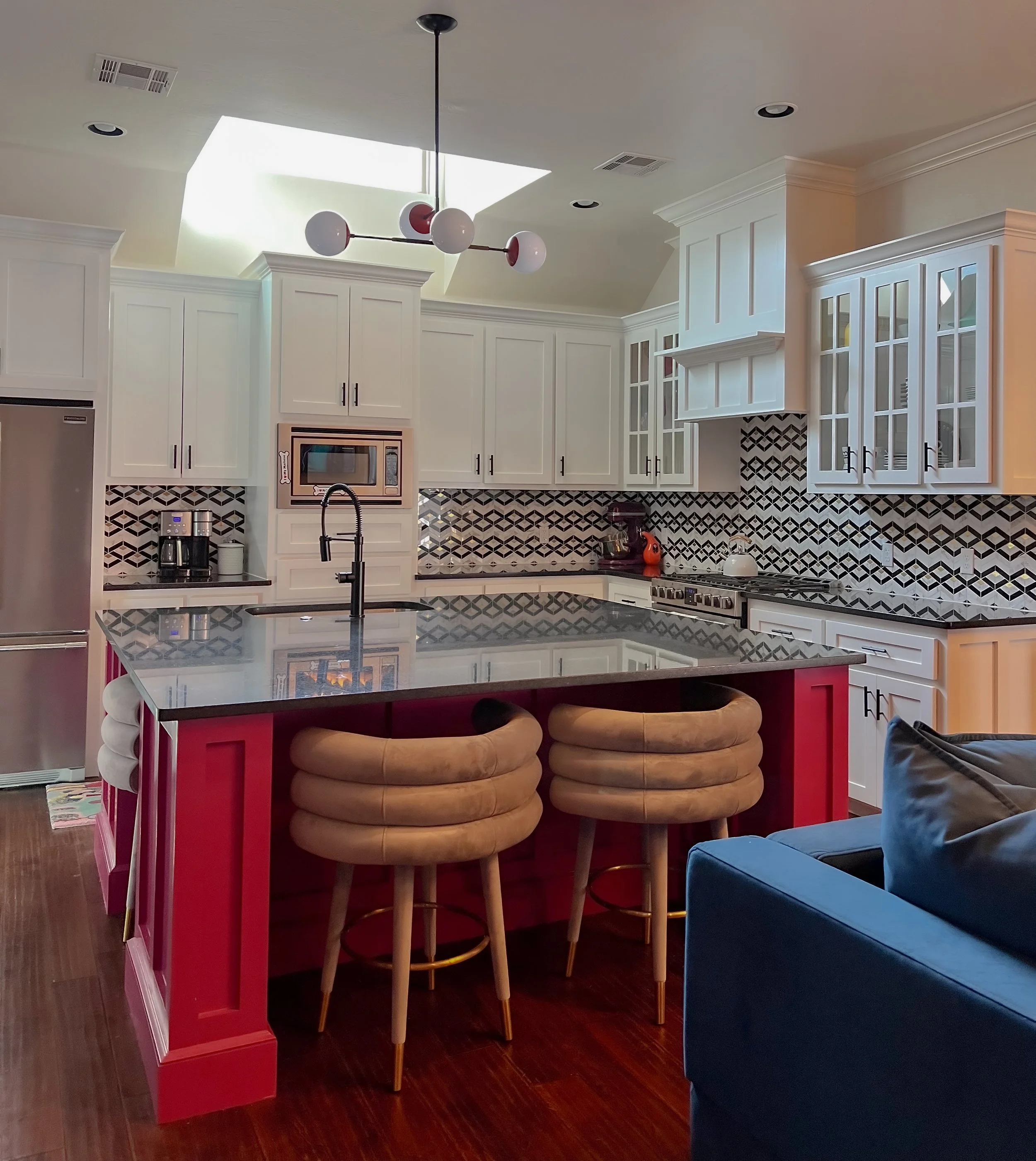 Modern kitchen with white cabinetry, black and white patterned backsplash, central island with a black countertop, and three tan cushioned barstools. A skylight and ceiling light fixture are also visible.