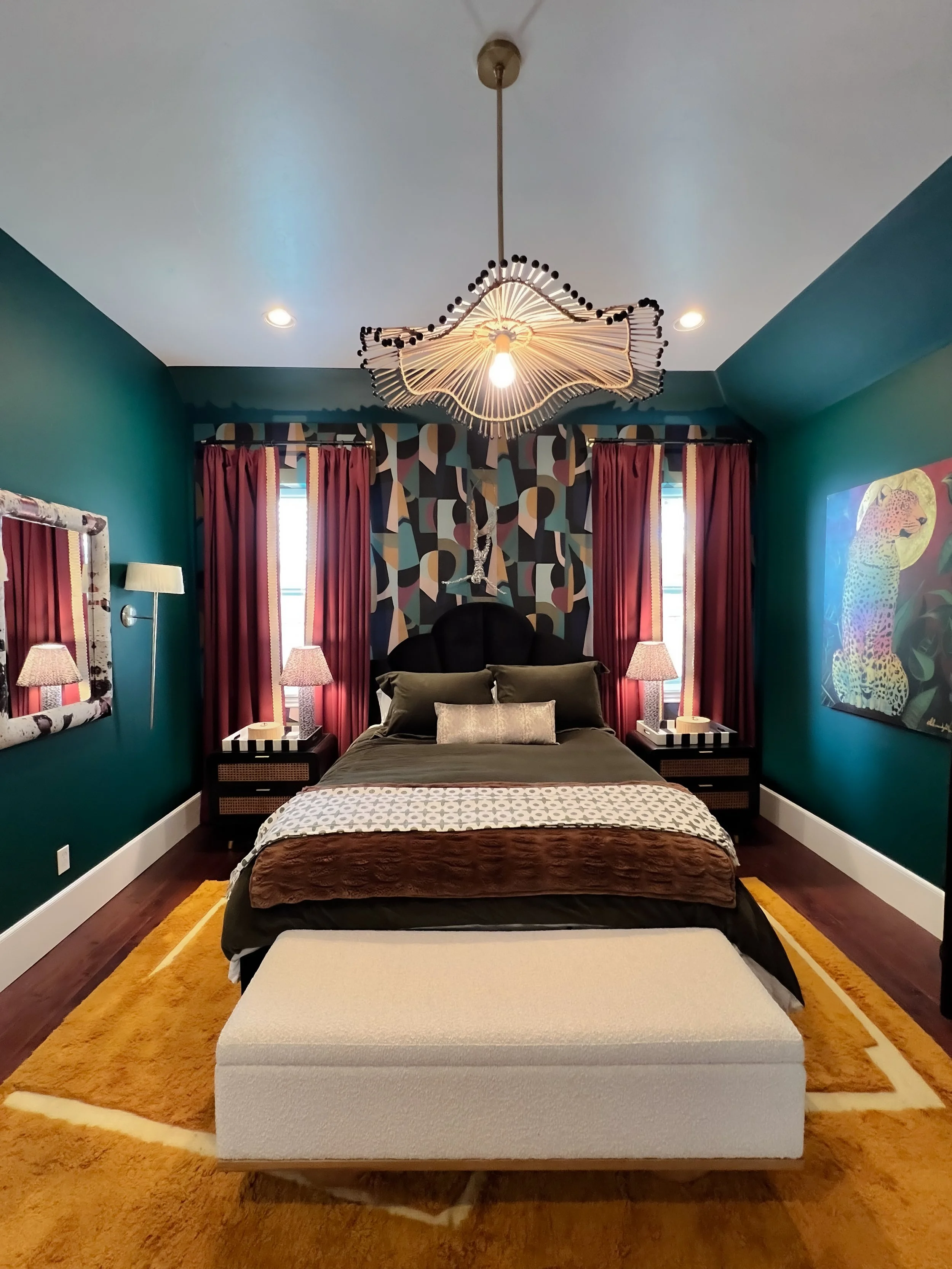 A bedroom with dark green walls, a black headboard, and colorful geometric wallpaper behind the bed. There are two windows with red curtains, a chandelier hanging from the white ceiling, and a large framed painting of a leopard on the right wall. The