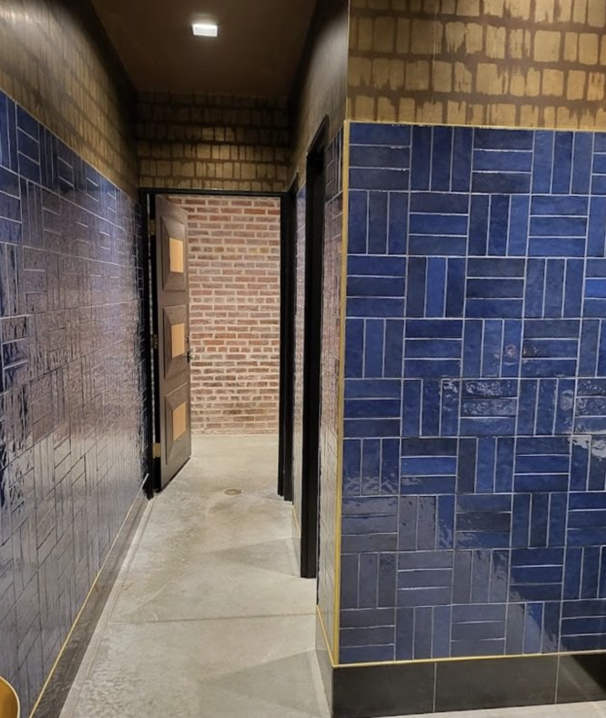 A corridor with brick walls and ceramic tiled walls, one door slightly open leading to another room with brick walls.