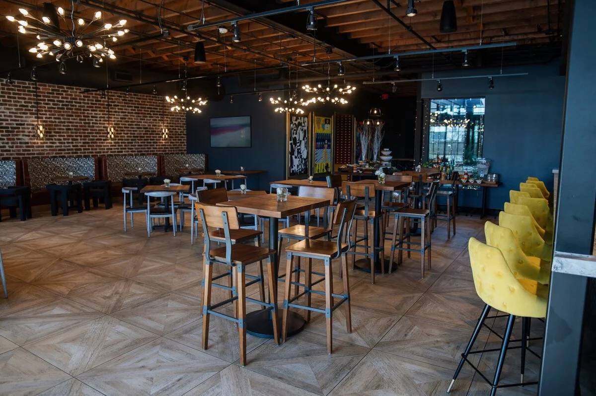 Modern restaurant interior with wooden tables and chairs, yellow bar stools, exposed brick wall, decorative lighting fixtures, and large windows.