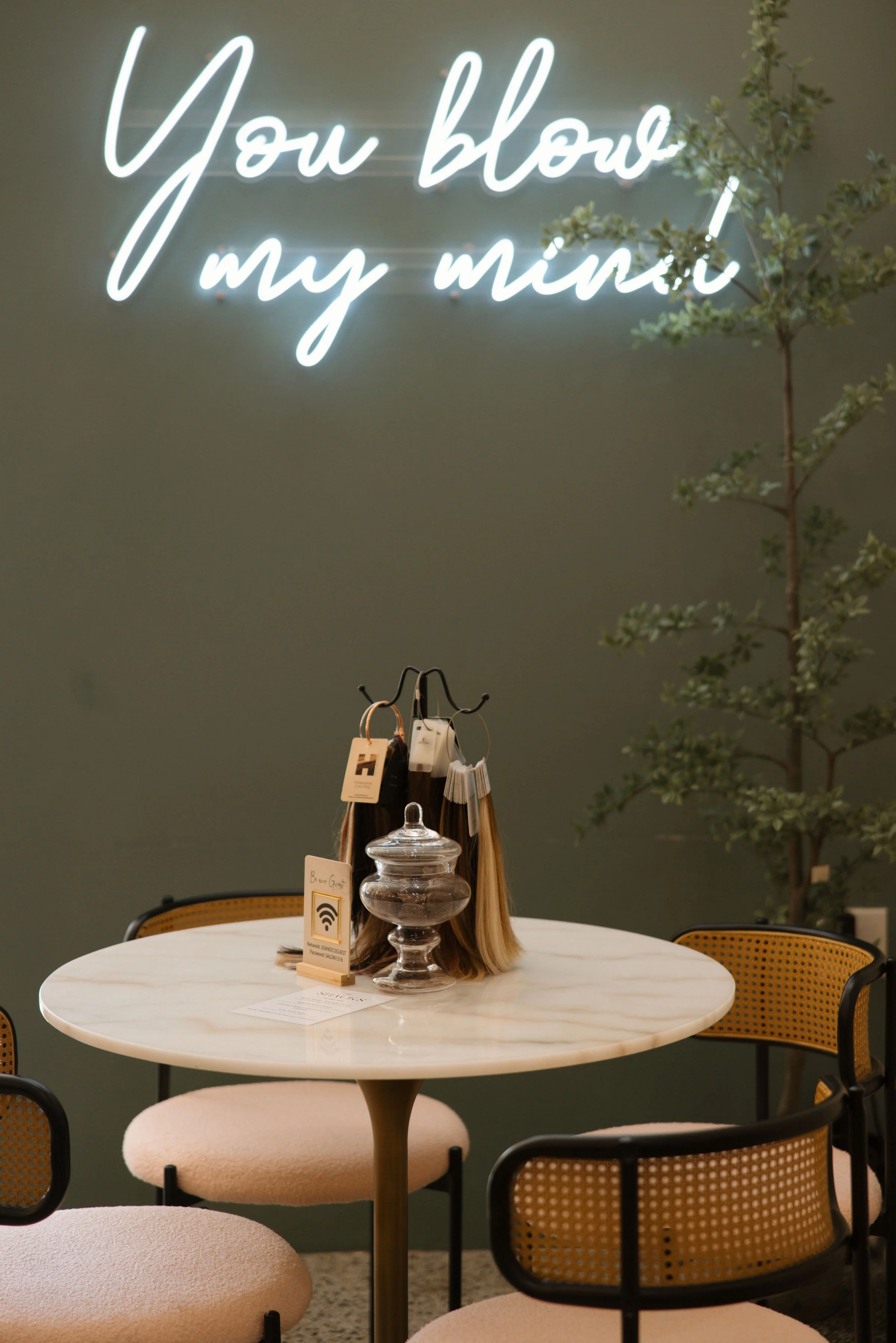 A round marble table with four beige cushioned chairs in a cafe. On the table, there is a glass jar and a small stand with a Wi-Fi sign. A hanging hair dye or sample color display and a small plant are on the wall behind the table. A neon sign on the