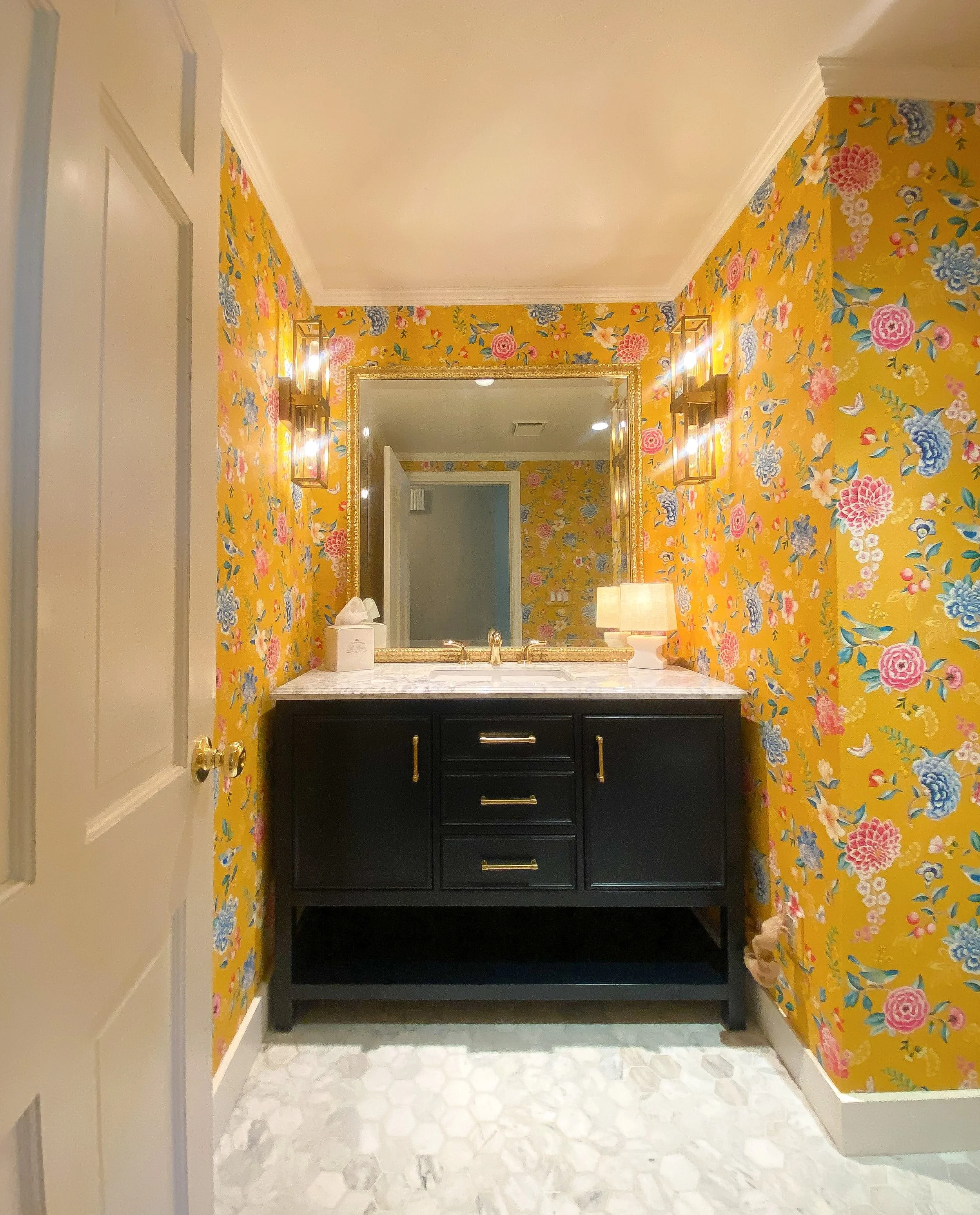 Bathroom vanity with a dark cabinet, marble countertop, large mirror, tissue box, and a lamp, surrounded by yellow floral wallpaper.