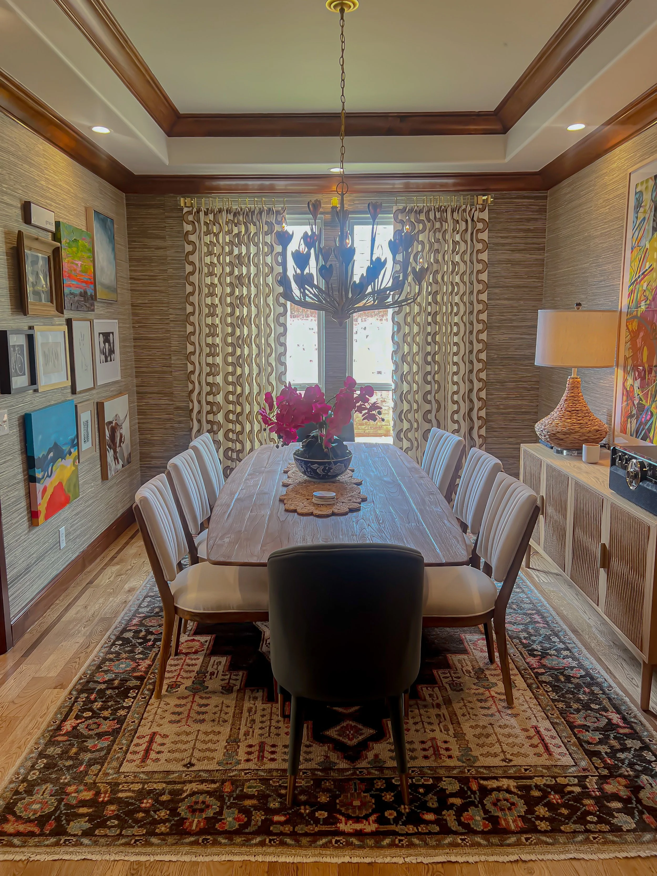 A dining room with a wooden table surrounded by eight chairs, a floral centerpiece, art on the walls, a chandelier, curtains, a sideboard with a lamp, and a patterned rug.