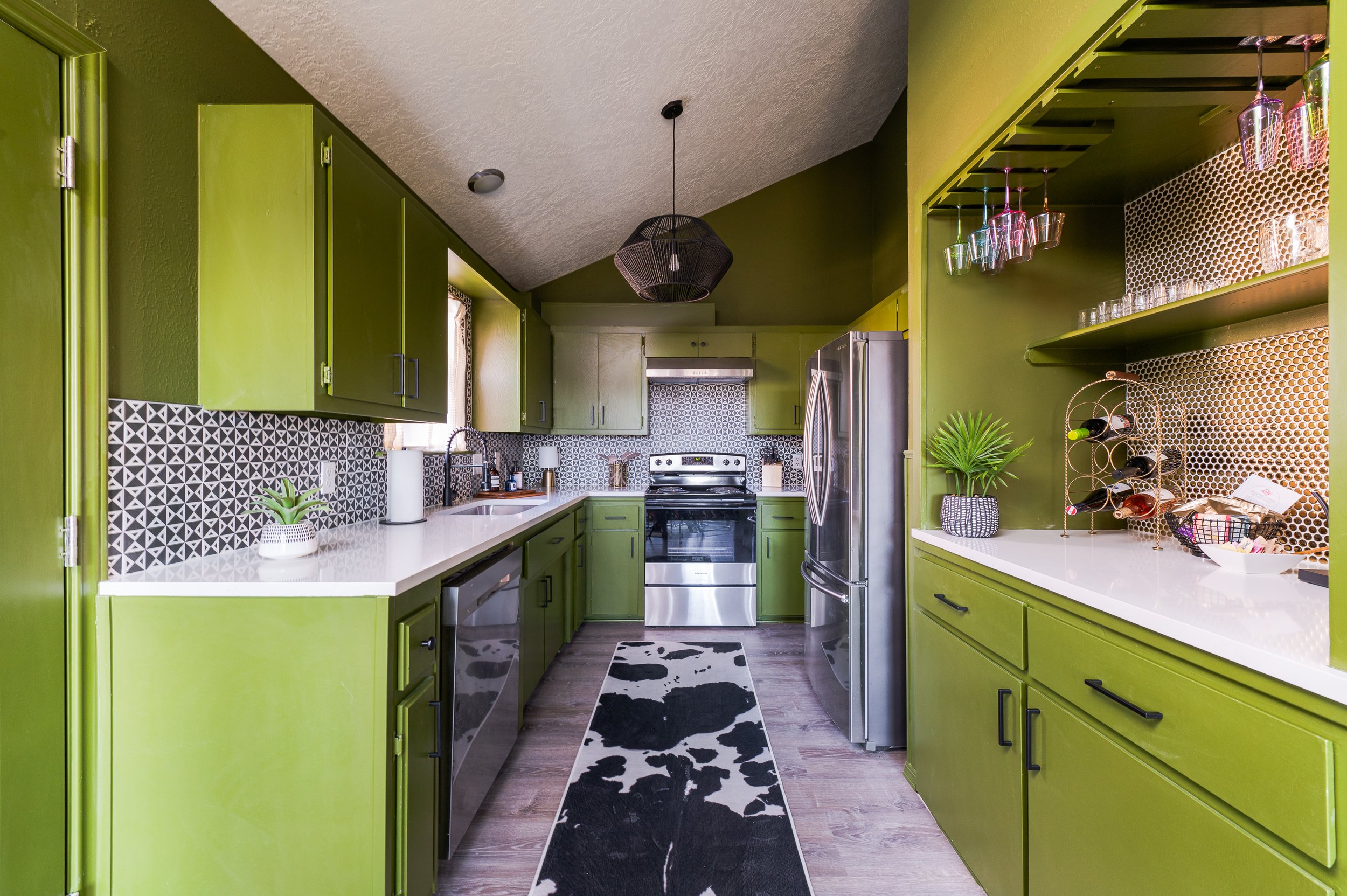 Bright kitchen with lime green cabinets, black and white geometric backsplash, stainless steel appliances including dishwasher, oven, and refrigerator. Black cowhide rug on wooden floor, small plants on countertops, and hanging glassware above a whit
