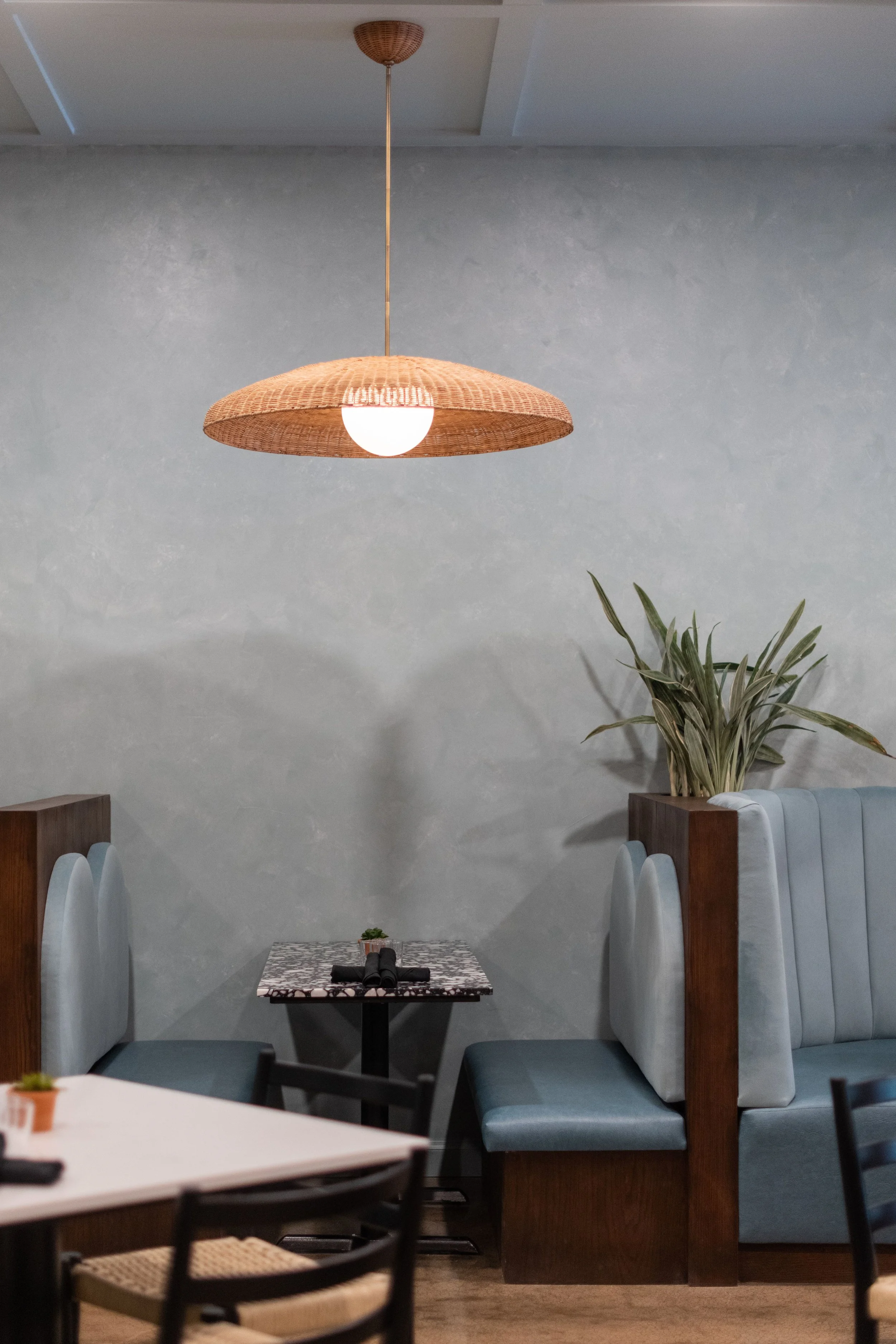 Interior of a restaurant or cafe with a hanging wicker pendant light, a small table with binoculars and a plant, and a booth seat with a tall plant behind it.