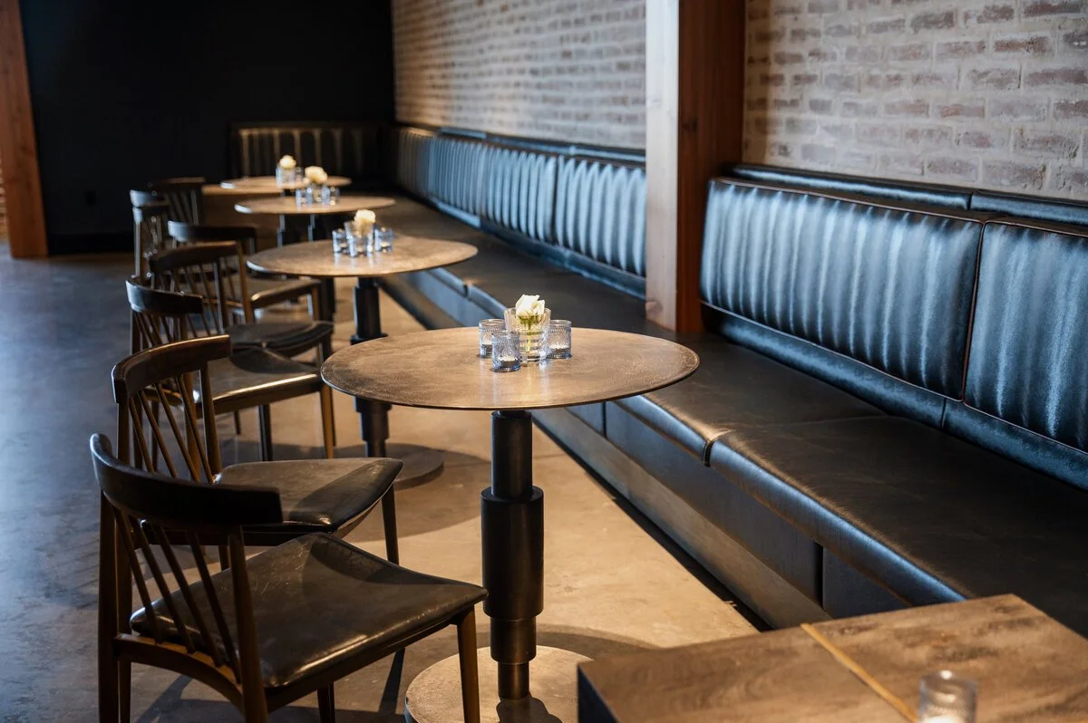 Empty restaurant dining area with round wooden tables, black chairs, a long black bench, and small flower arrangements on the tables.