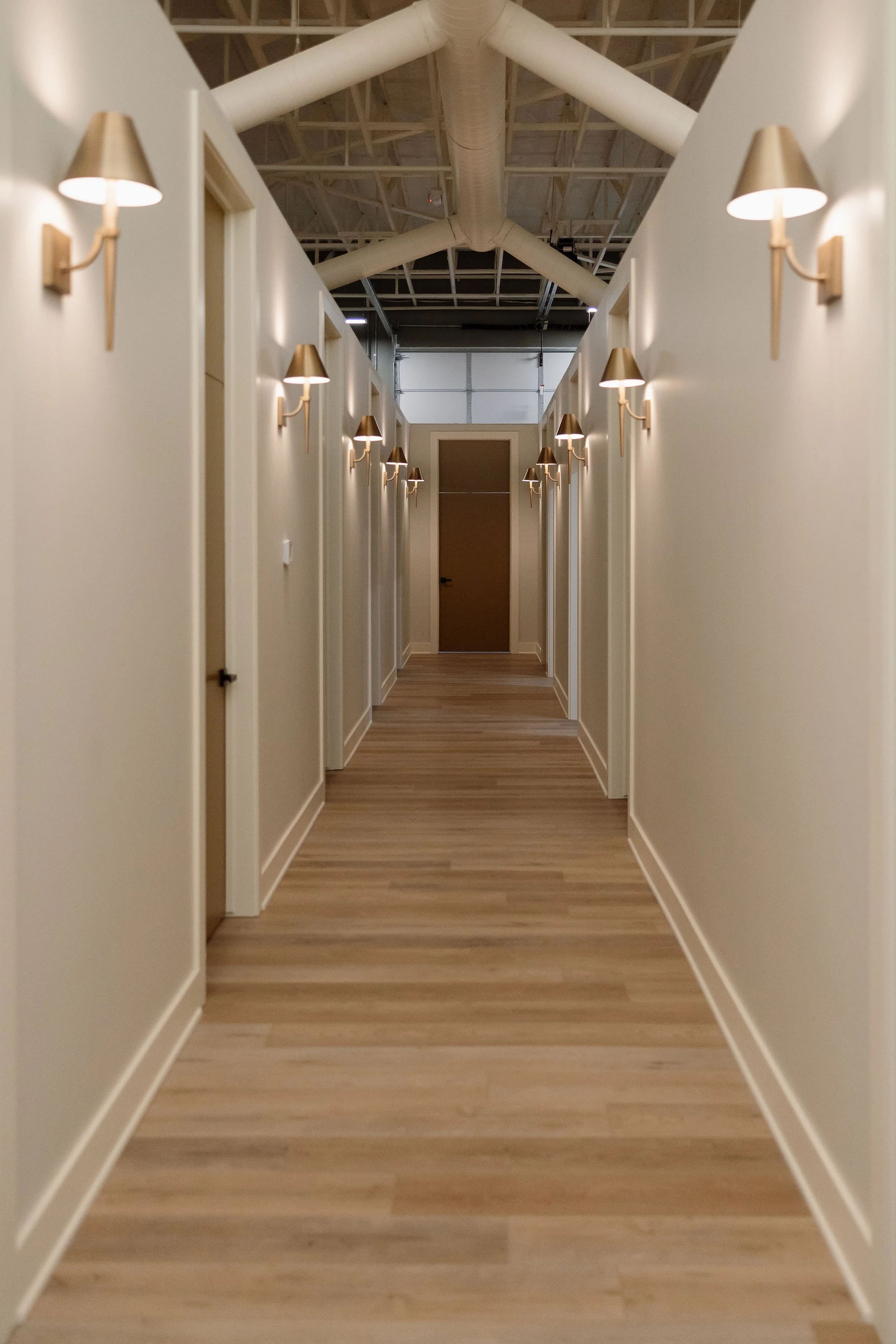 Empty hallway with beige walls, wooden flooring, and wall-mounted lamps on both sides, leading to a door at the end.