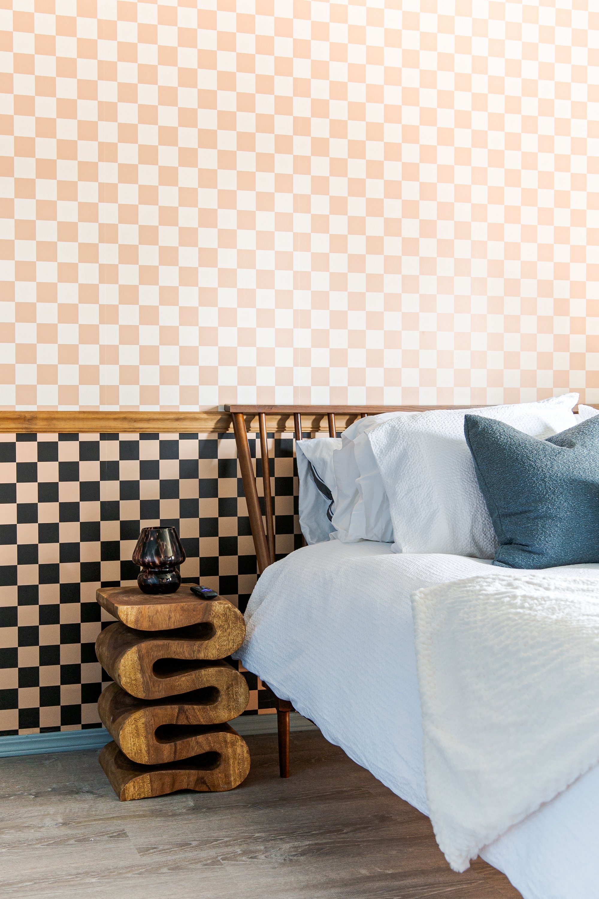 A bedroom with a black and beige checkered headboard, white bedding, and a dark gray pillow, next to a uniquely designed wooden nightstand with a black lamp and remote, with a pink and beige checkered accent wall.