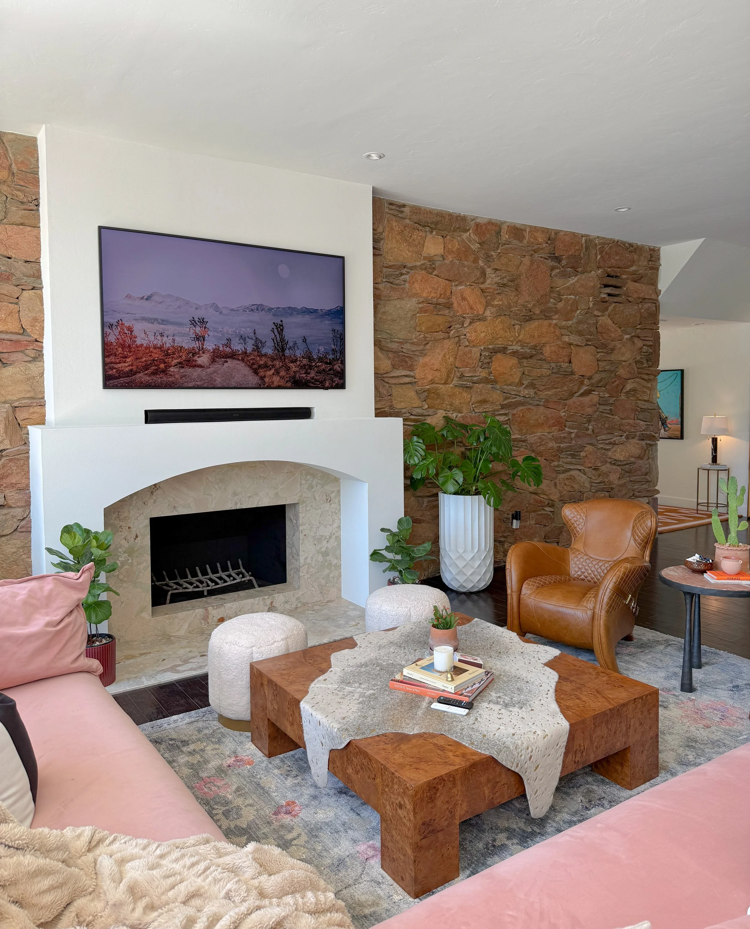 Living room with brick and white fireplace, flat-screen TV, and leather armchair, with plants, a wooden coffee table, and decorative items.
