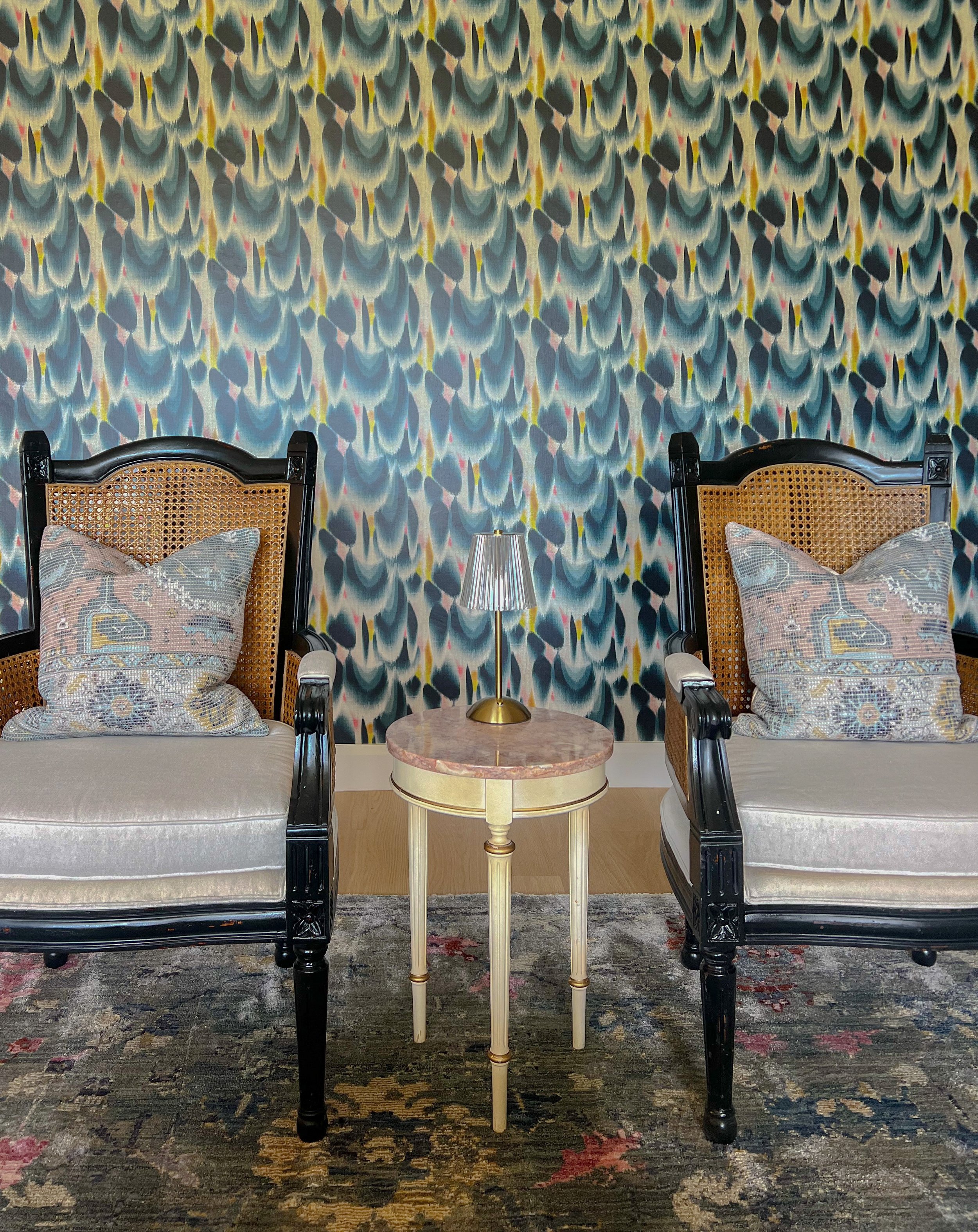 Two armchairs with black frames and caned backs, each with a decorative pillow, are positioned on either side of a small round side table with a marble top. A small lamp with a striped shade sits on the table. There is a colorful, abstract patterned 