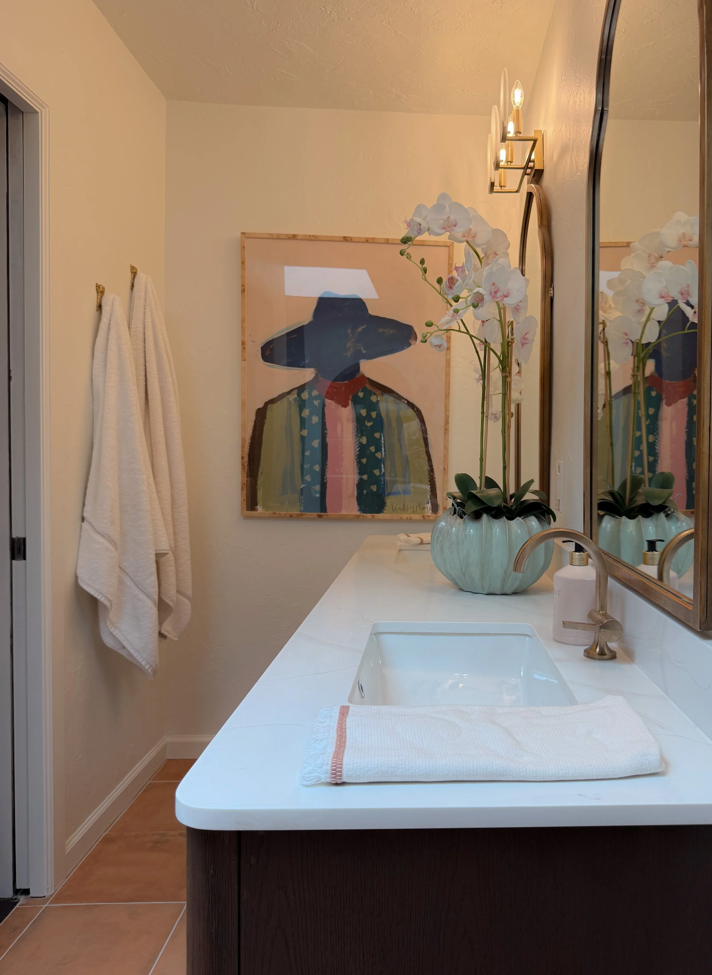 A bathroom with a white countertop sink, a large mirror, a vase with orchids, a framed painting of a person wearing a hat and a jacket, and wall-mounted light fixtures.