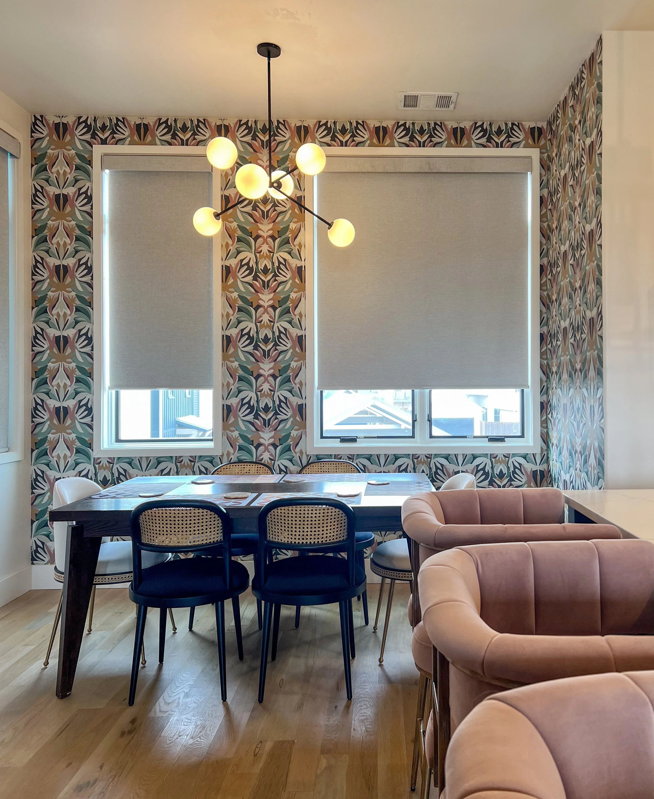 Dining room with patterned wallpaper, wooden table, black and white chairs, plush pink armchairs, large windows with blinds, and a modern light fixture.