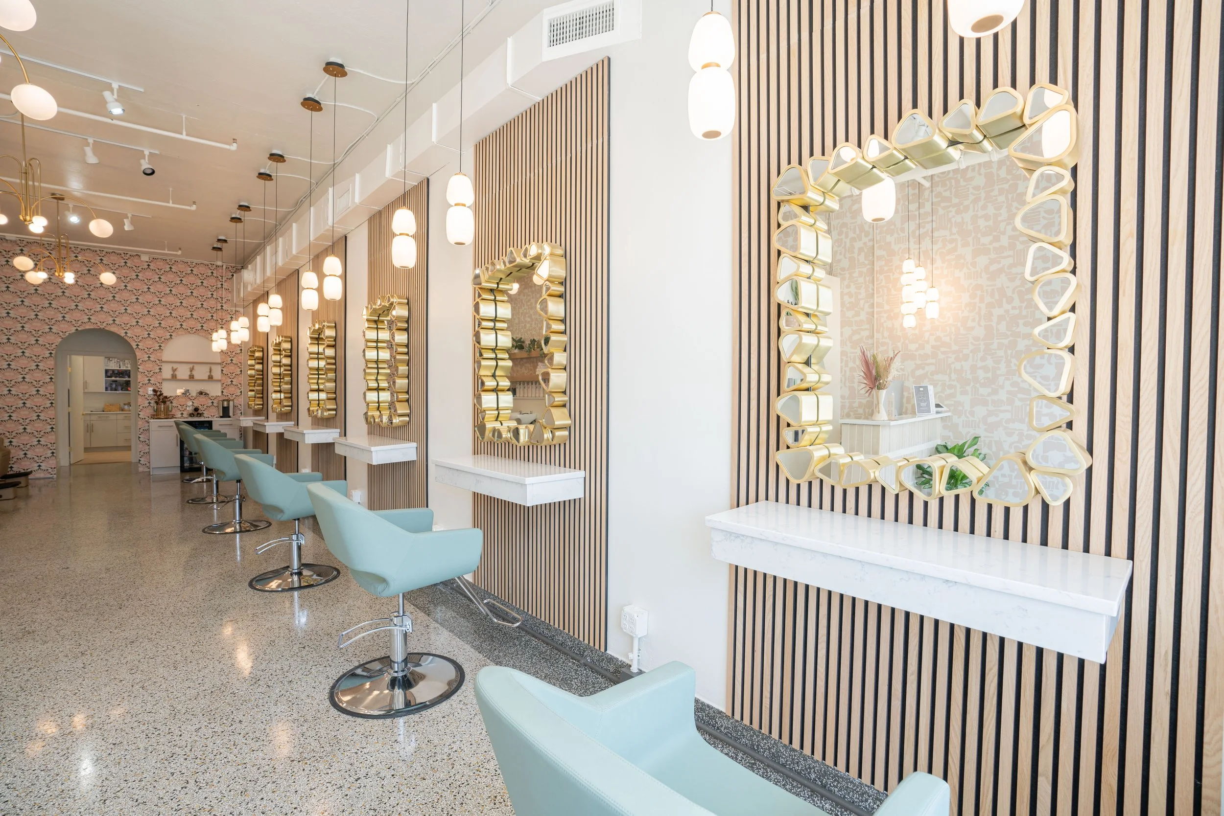 A row of salon chairs and mirrors in a modern hair salon with pastel color decor and hanging lights.