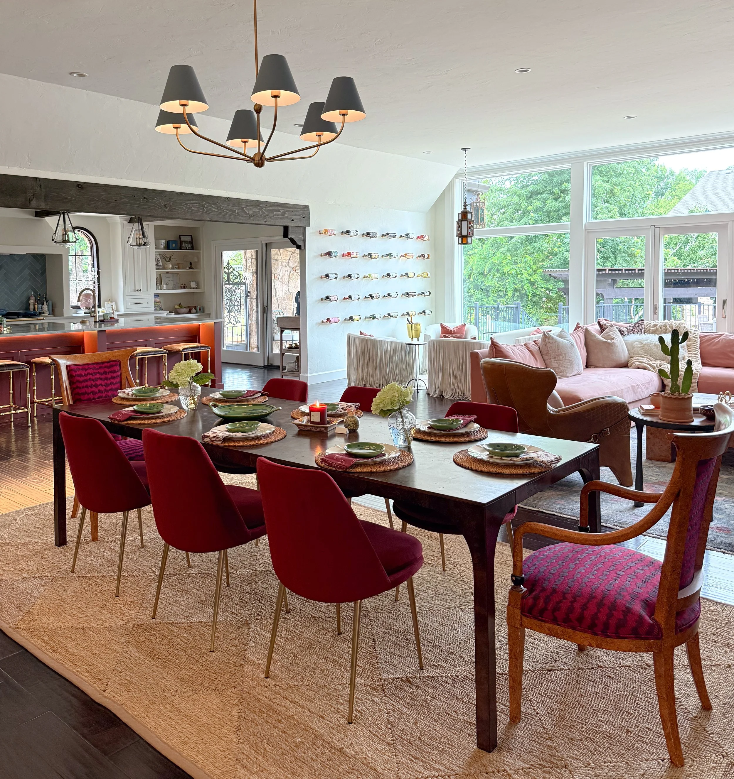 A dining room with a long wooden table set with green dishes and woven placemats, surrounded by red and wooden chairs, leading into a cozy living room with pink and brown sofas, a cactus plant, and large windows showing a green outdoor area.