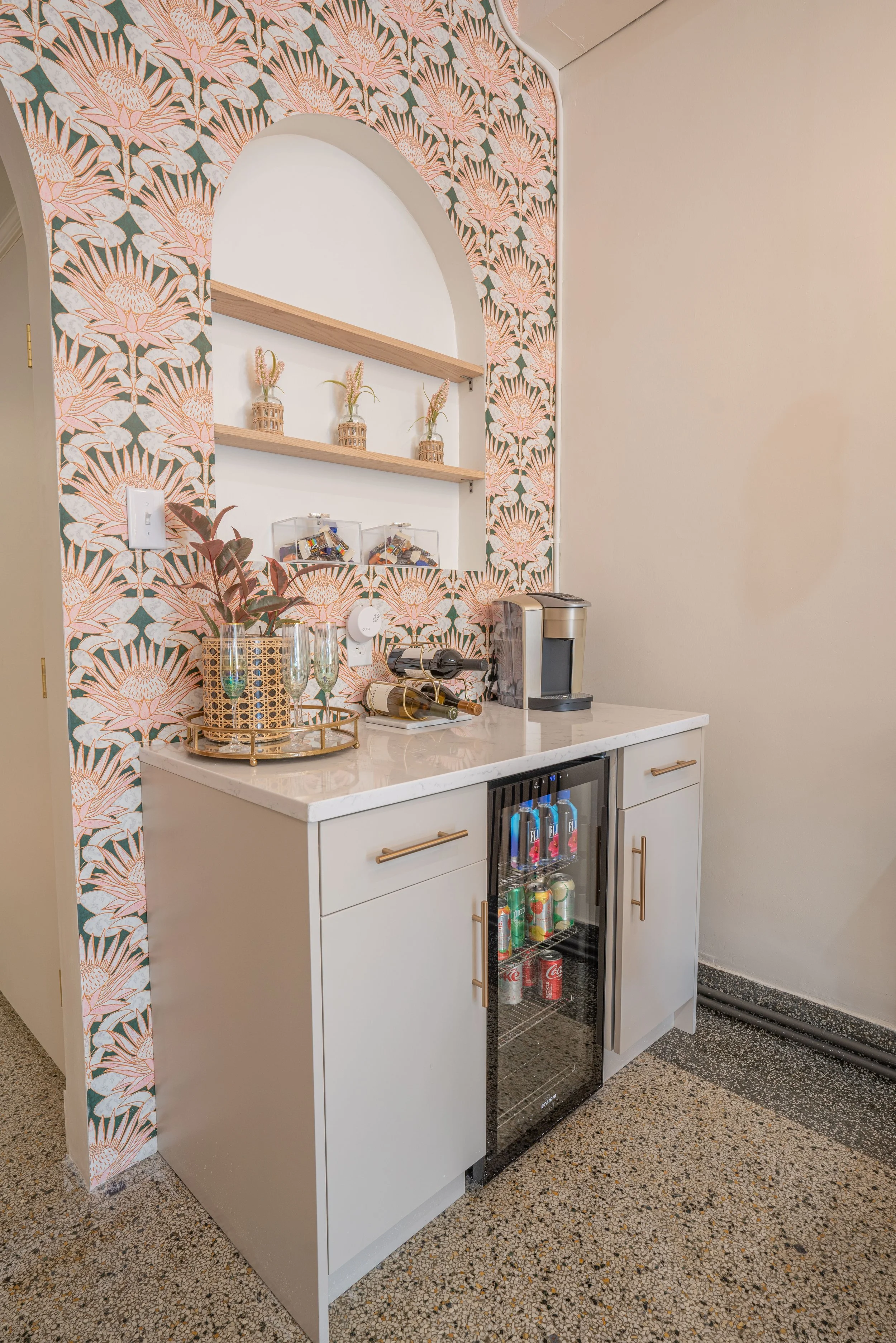 A small bar area with a white cabinet, a built-in mini fridge stocked with soda cans and bottled water, and a white countertop. There are decorative items including a tray with glasses, a wine bottle on a corkscrew, and a coffee machine. The wall beh