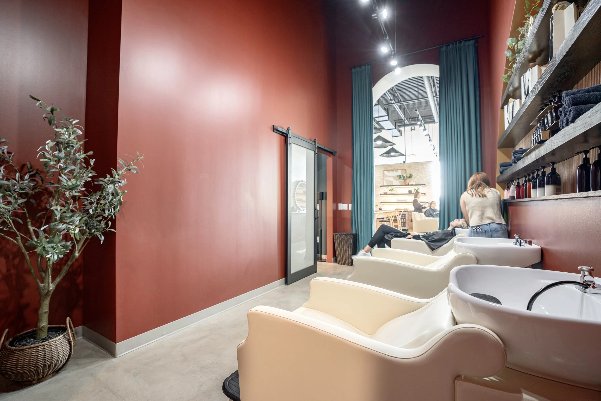 Inside a modern hair salon with red walls, white shampoo sinks, and shelves with hair products. Two women are sitting in chairs, one getting her hair washed, and the salon has a high ceiling with blue curtains and decorative plants.