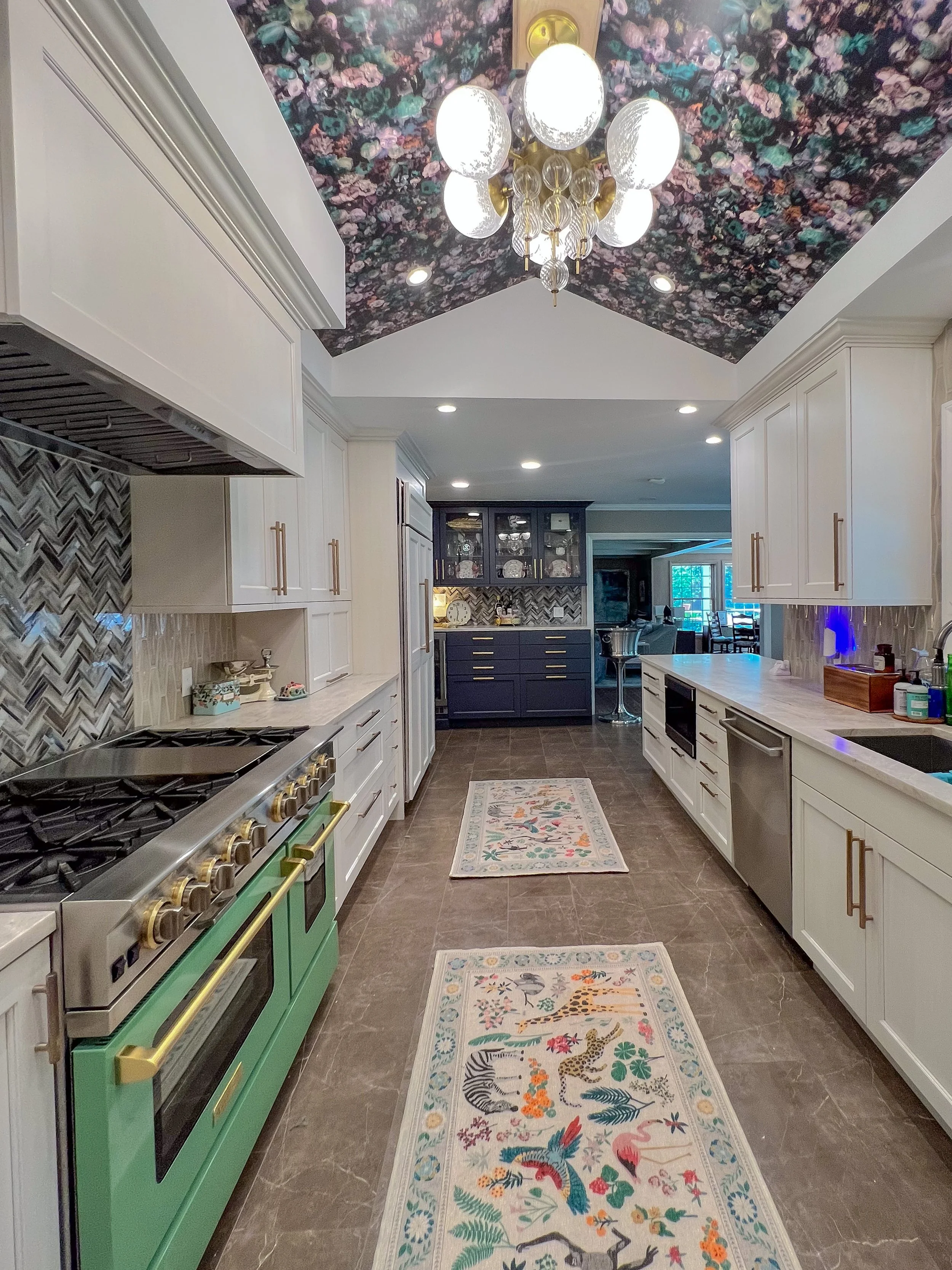 A modern kitchen with white cabinets, a green oven, and a colorful floral and animal-themed rug on a brown tiled floor. The ceiling has floral wallpaper and a chandelier with five glass globes.