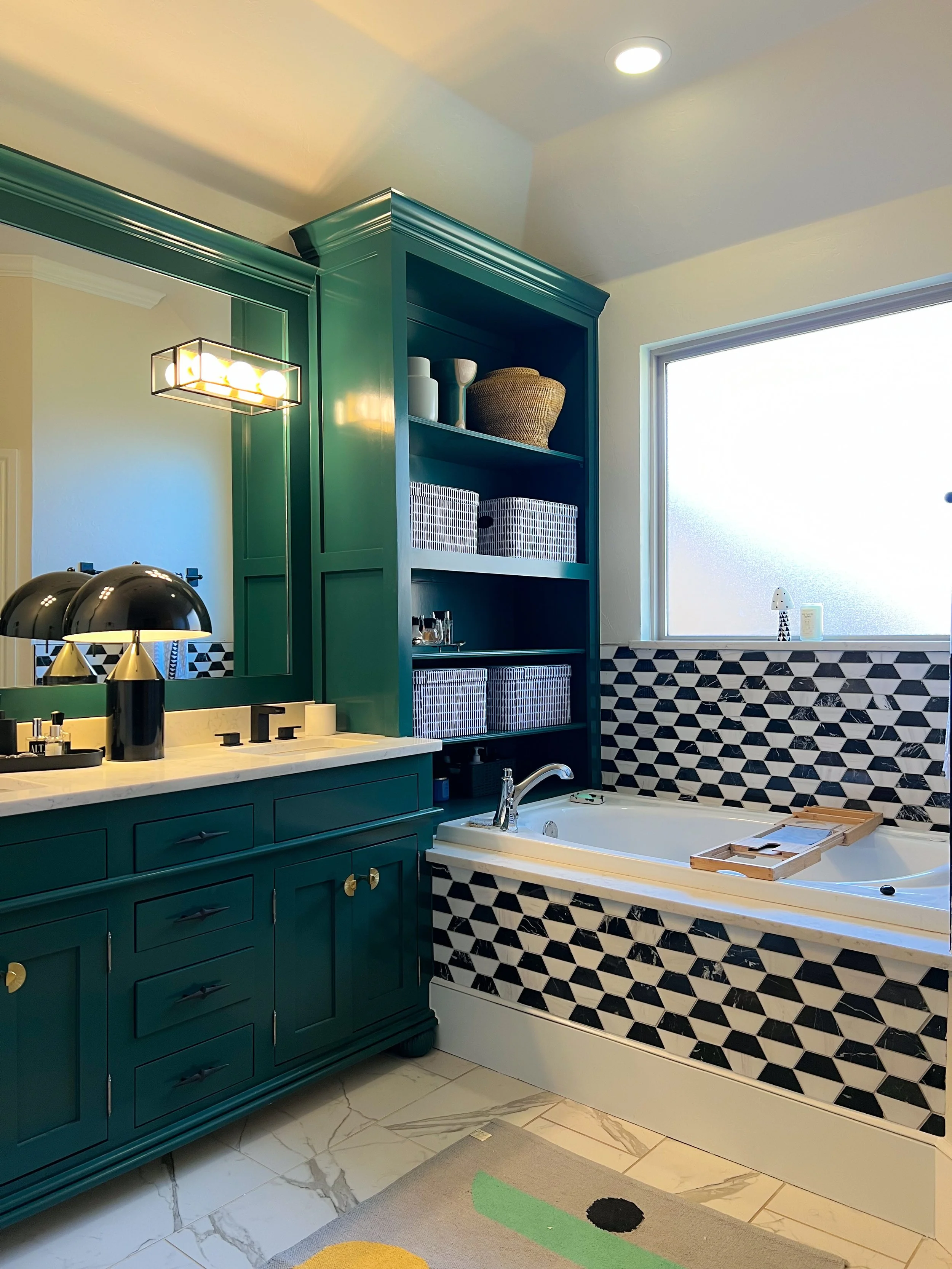 A bathroom with a green vanity, a bathtub with black and white geometric tile, a large mirror, open shelving with baskets and decor, and a window letting in natural light.