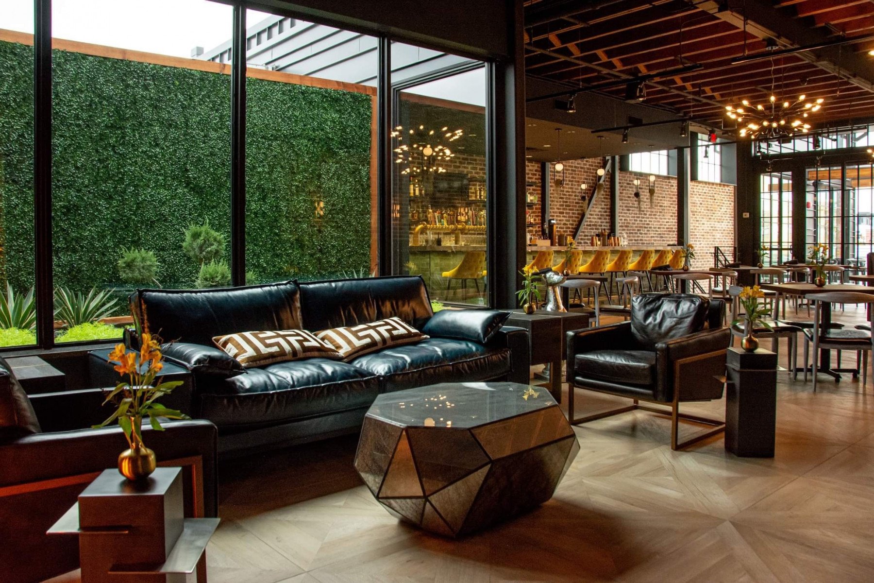 Modern lounge area with black leather sofas, patterned pillows, and a geometric coffee table. Large windows reveal a green outdoor wall. In the background, a bar with yellow chairs, wooden tables, and ambient lighting create a cozy atmosphere.