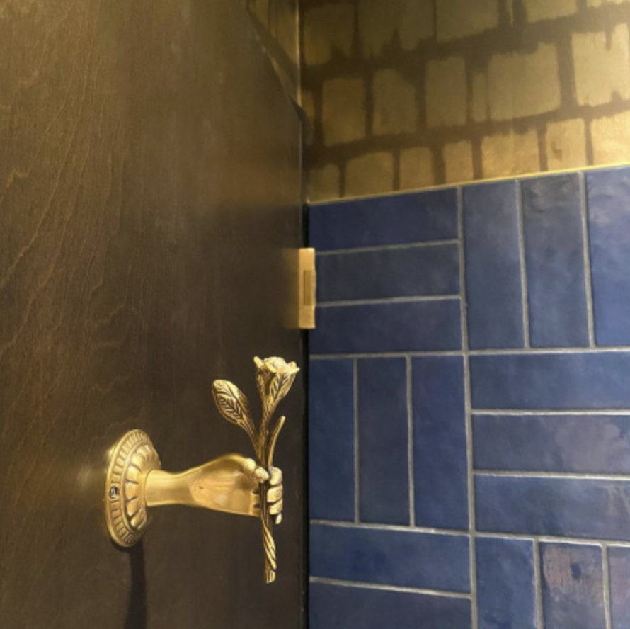 A small gold-colored candlestick holder shaped like a hand holding a rose, placed on a dark table against a background of blue tiles and brick wall.