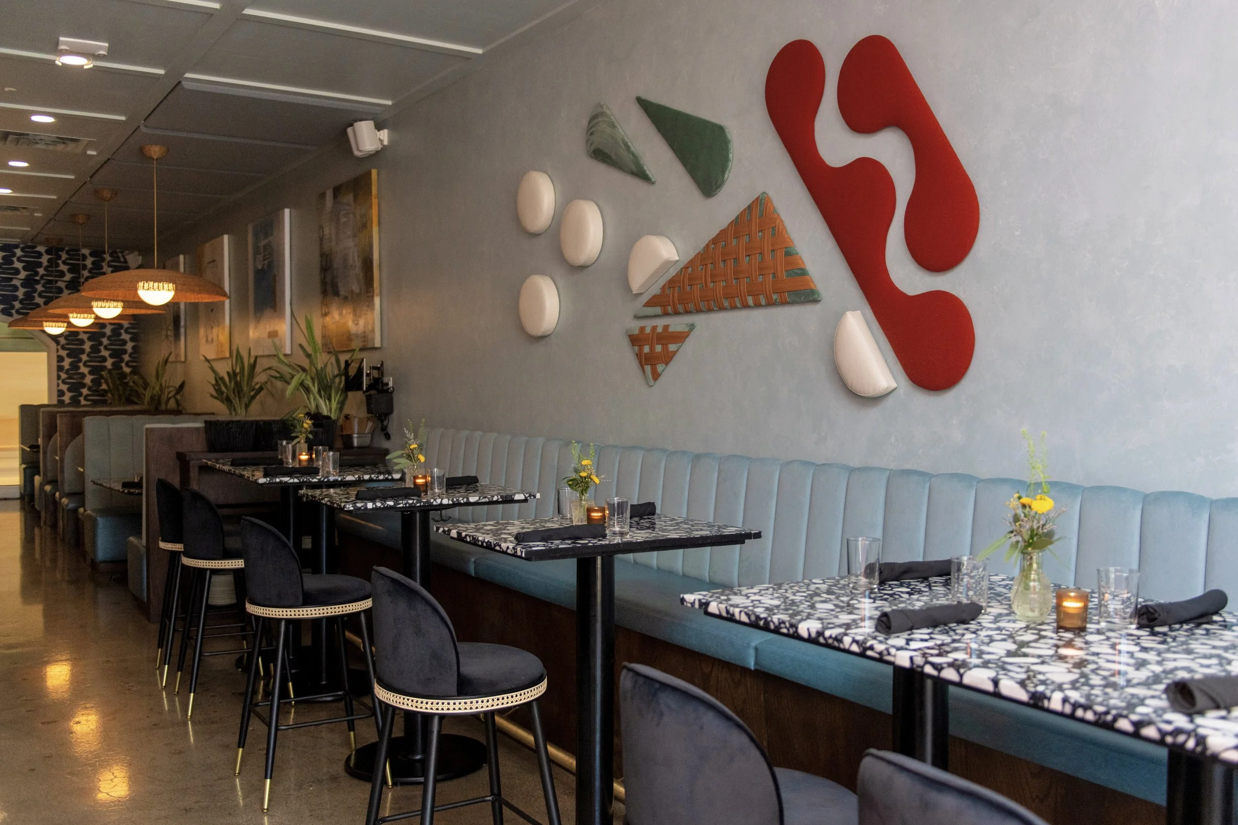 Interior of a modern restaurant with a row of tables, black chairs, and a light blue upholstered bench along the wall. The tables are decorated with small vases of flowers and candles. The wall features abstract art with various shapes and colors.