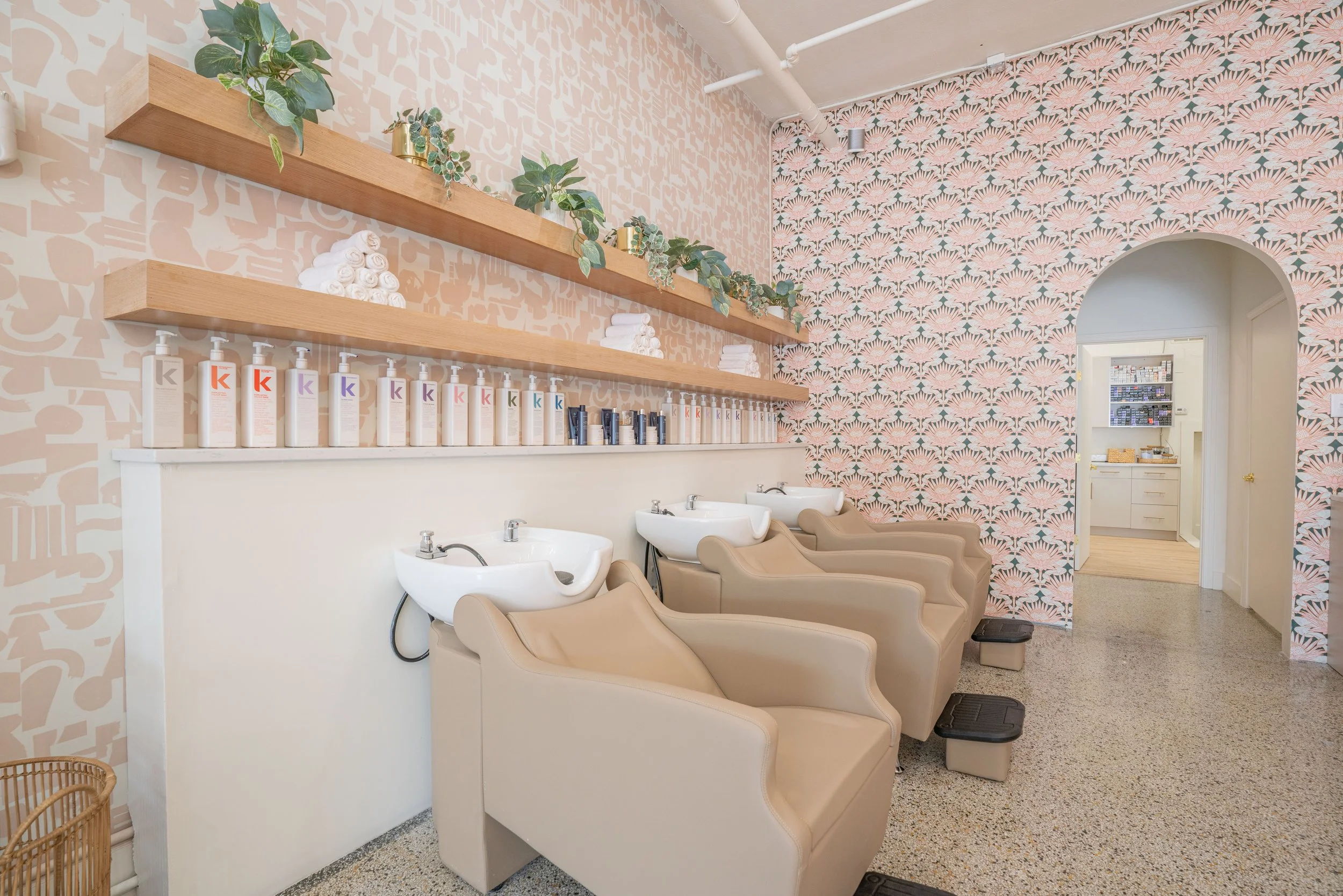 Interior of a hair salon or spa with three beige hair washing chairs and sinks, wooden shelves with plants and towels, patterned wallpaper, and a view into a small kitchen or reception area.